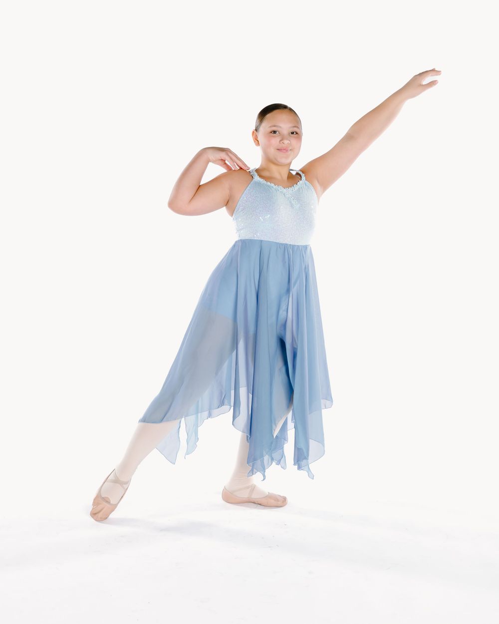 Dancer in flowing light blue dress and ballet shoes strikes graceful pose against white background.