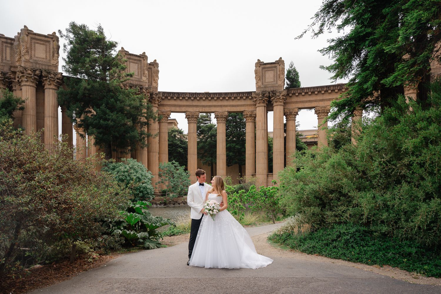 A couple in wedding attire stands beneath grand stone columns at the Palace of Fine Arts in San Francisco.