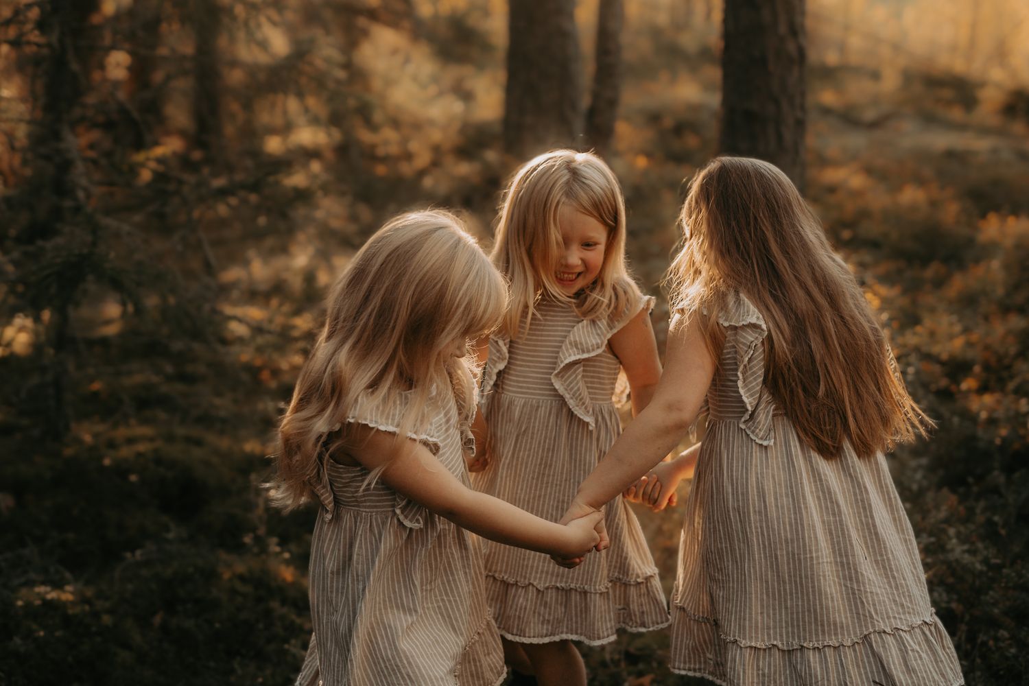 Three children in vintage style dresses share a joyful moment together in a sunlit forest during golden hour.