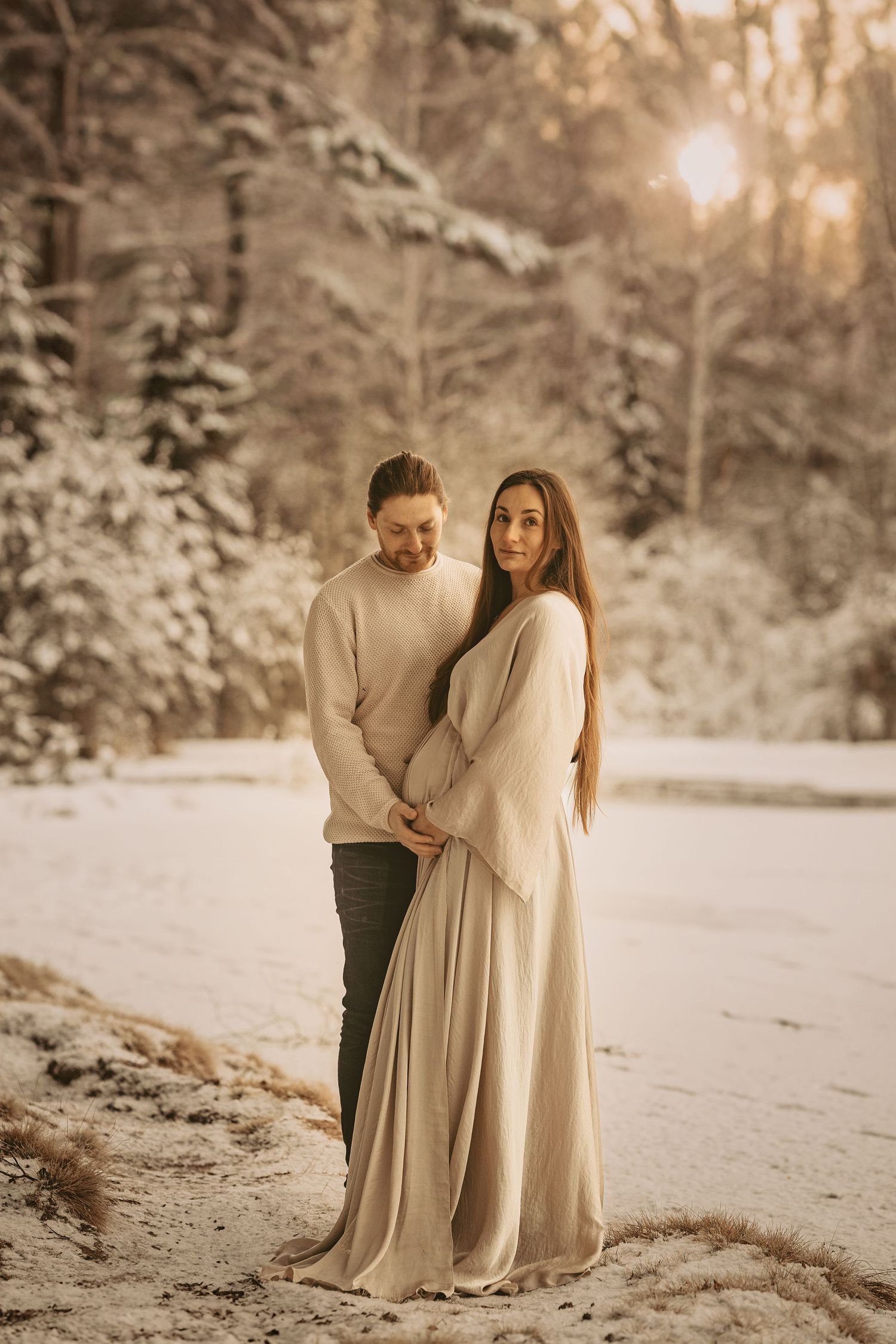 A couple in neutral-colored outfits embraces in a snowy forest during golden hour sunset.