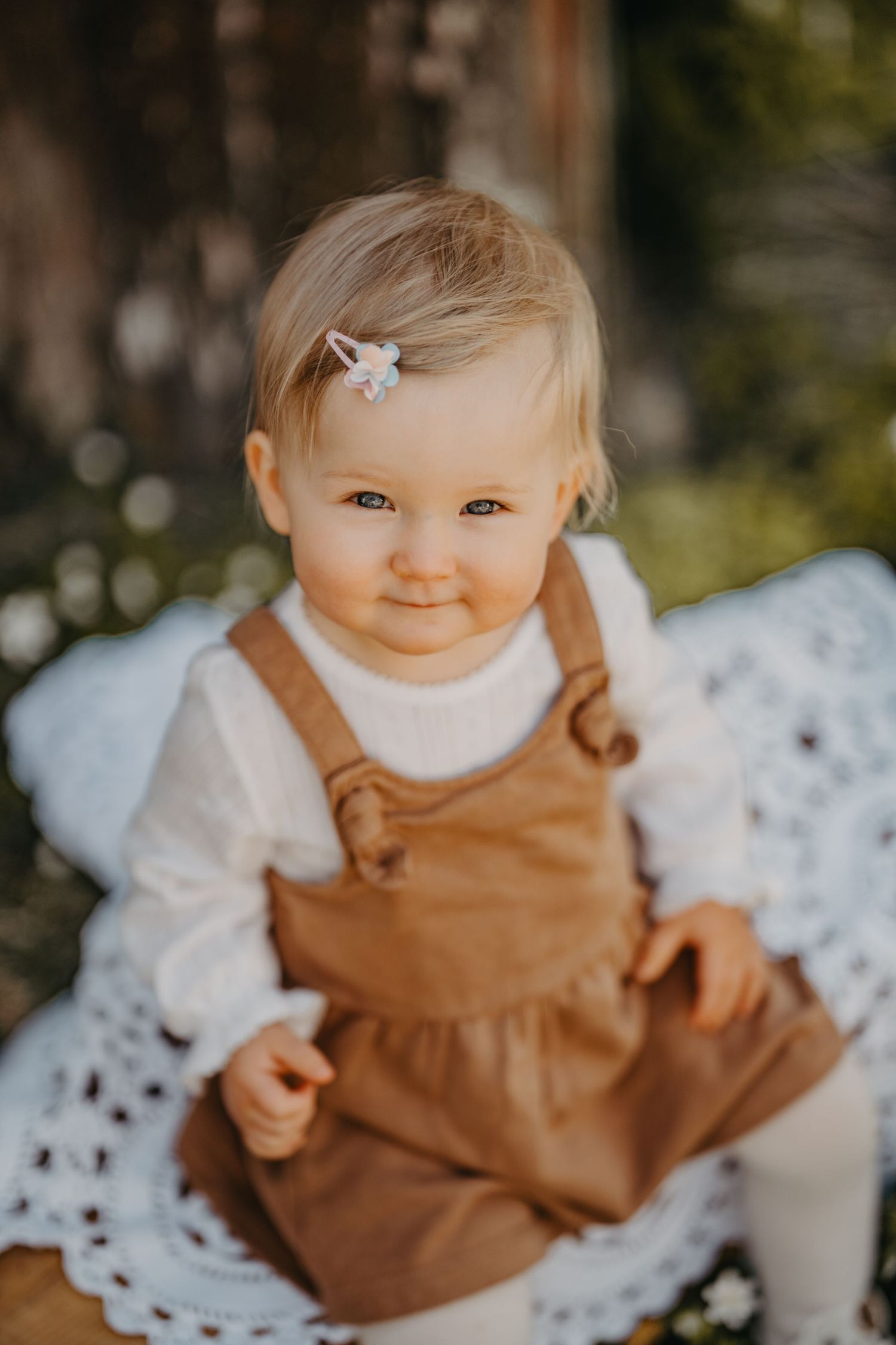 A baby wearing a brown pinafore dress and white sweater sits on a white lace blanket outdoors.