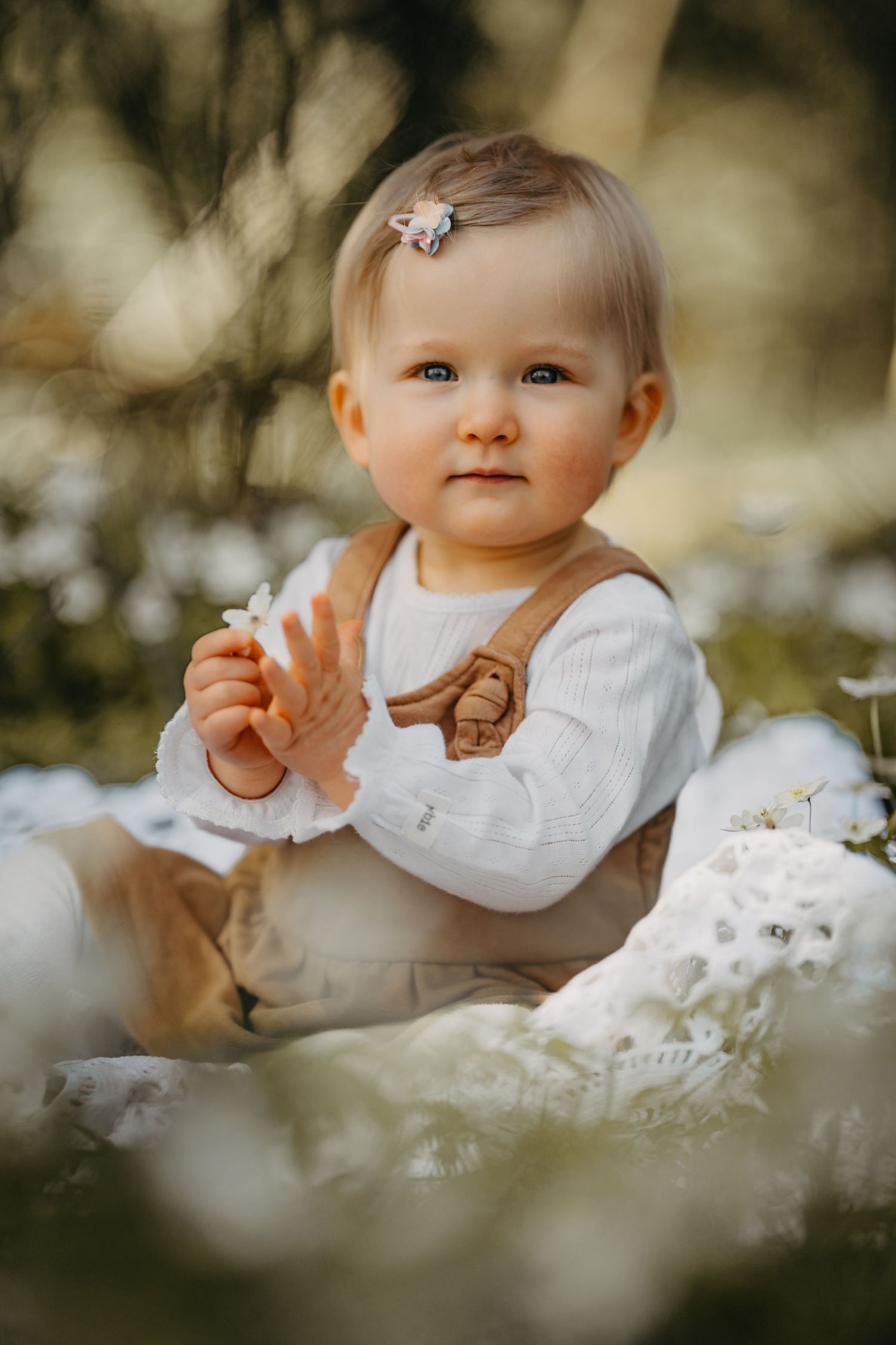 Baby wearing white sweater with brown design sits among soft bokeh background and white flowers.