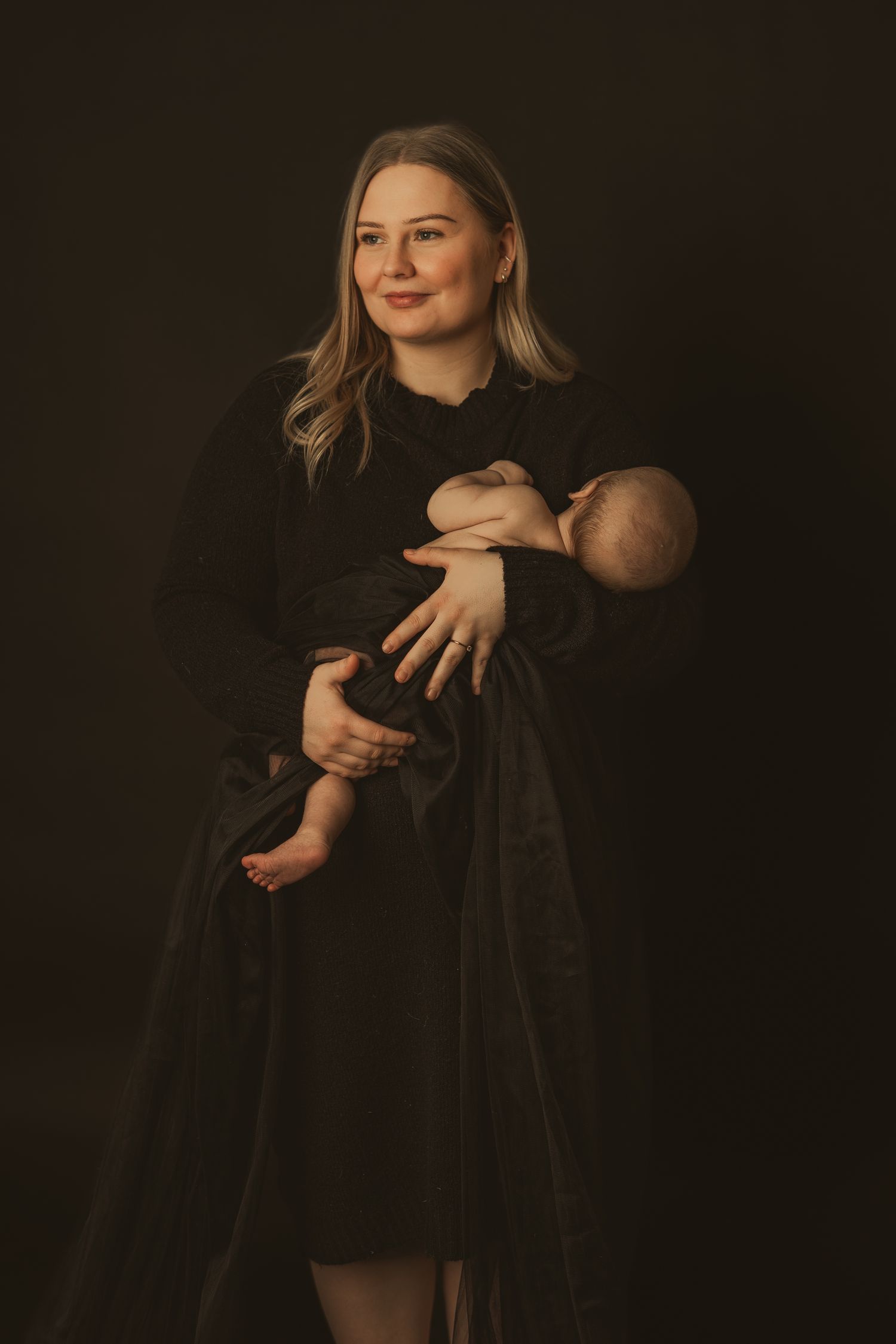 Studio portrait of a figure in a black dress cradling an infant against a dark background in moody lighting.