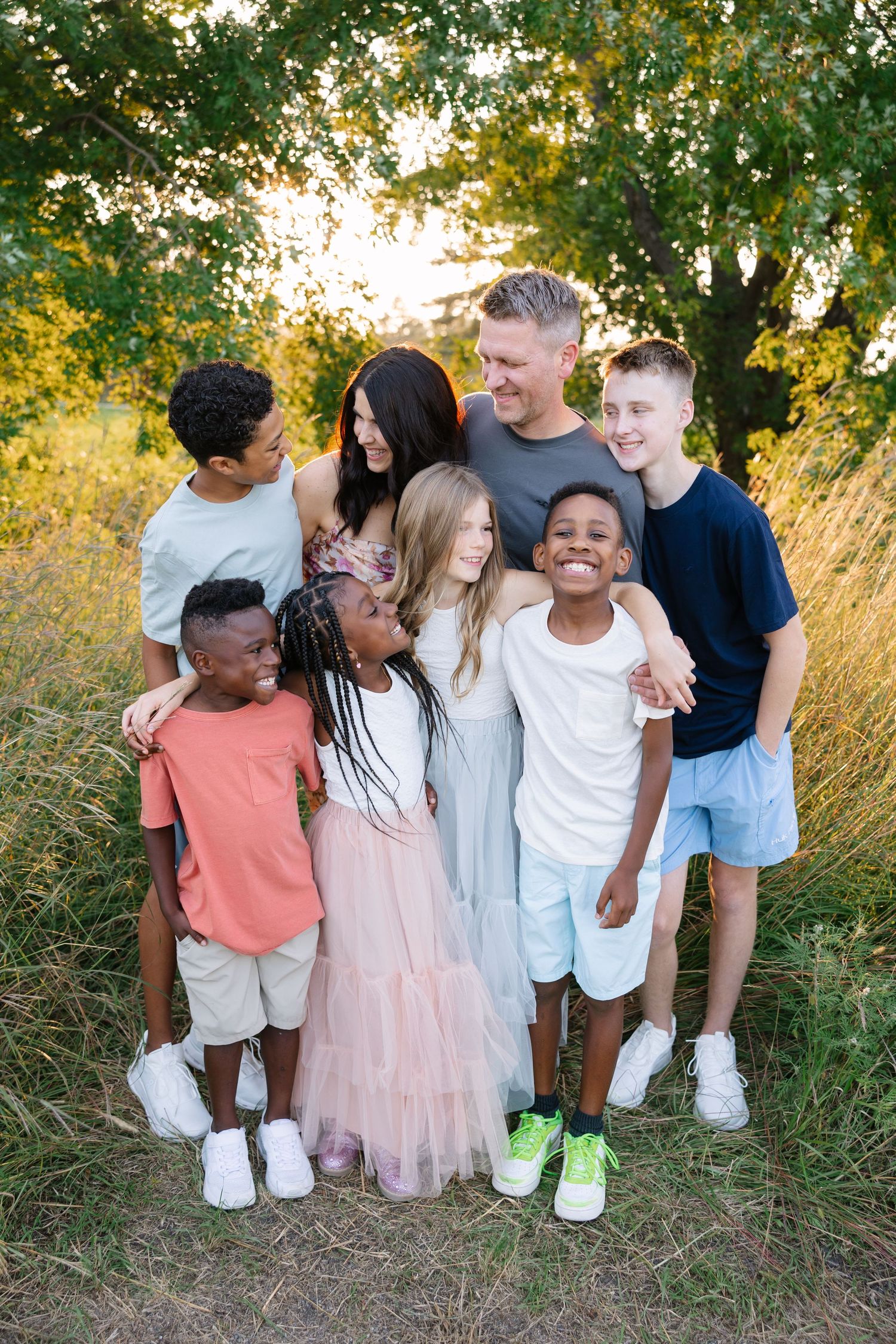 Happy family embraces and laughs together outdoors in a golden field at sunset amid lush green trees.