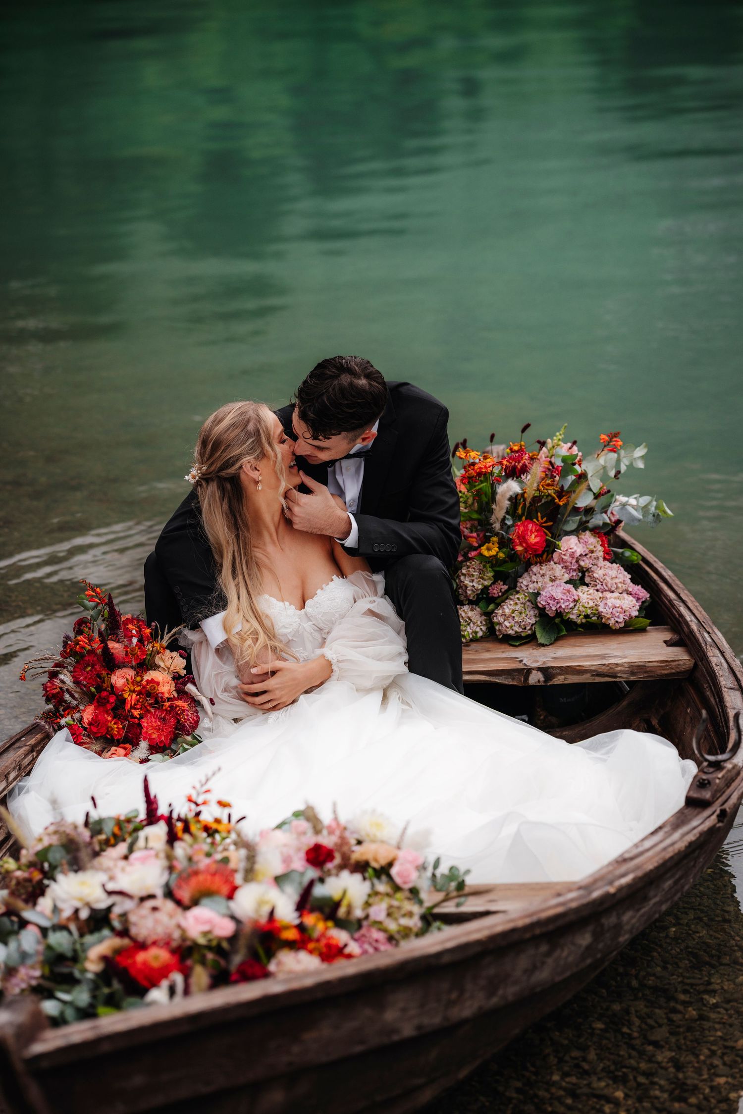 Newlywed couple sharing romantic kiss in wooden boat decorated with vibrant red and pink floral arrangements on calm water.