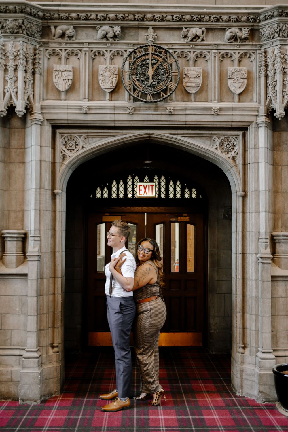 Elegant couple stands in ornate gothic archway with plaid carpet and decorative stonework at university or castle entrance.