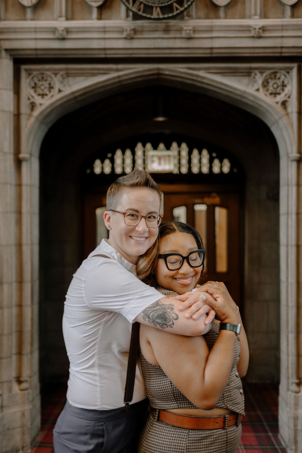 A loving couple embraces in an ornate archway while wearing glasses and retro-inspired outfits.
