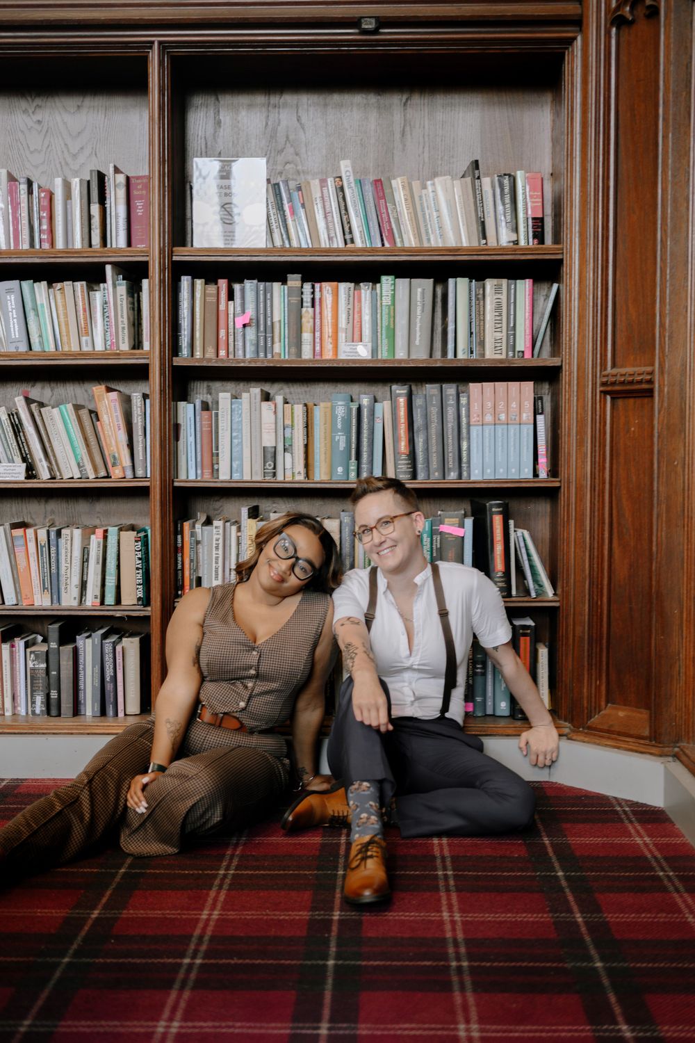 Two people relaxing on a plaid carpet against a library bookshelf in casual vintage-inspired outfits.
