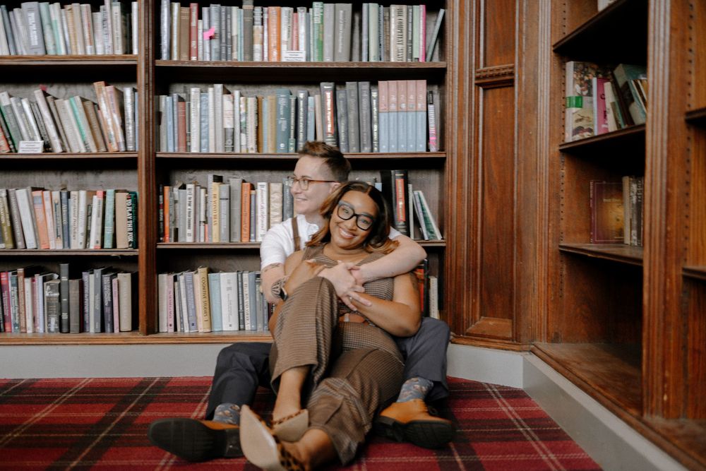 A couple relaxes together on a plaid blanket in a cozy library filled with wooden bookshelves.
