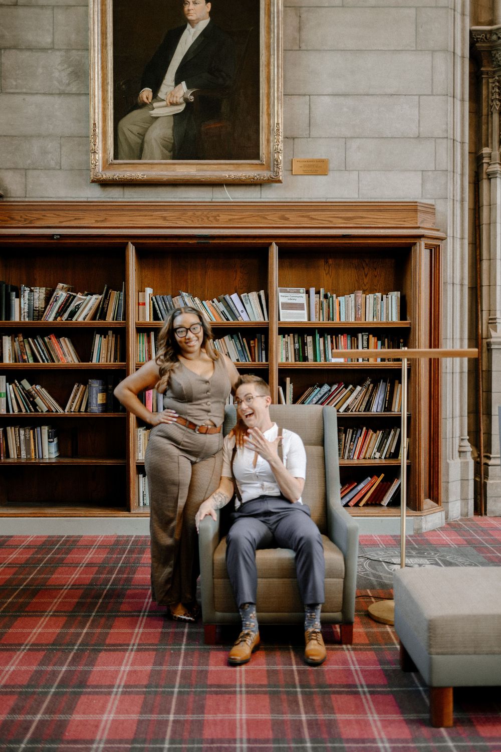 Two people share a playful moment in a library with plaid carpet and wooden bookshelves beneath a portrait painting.
