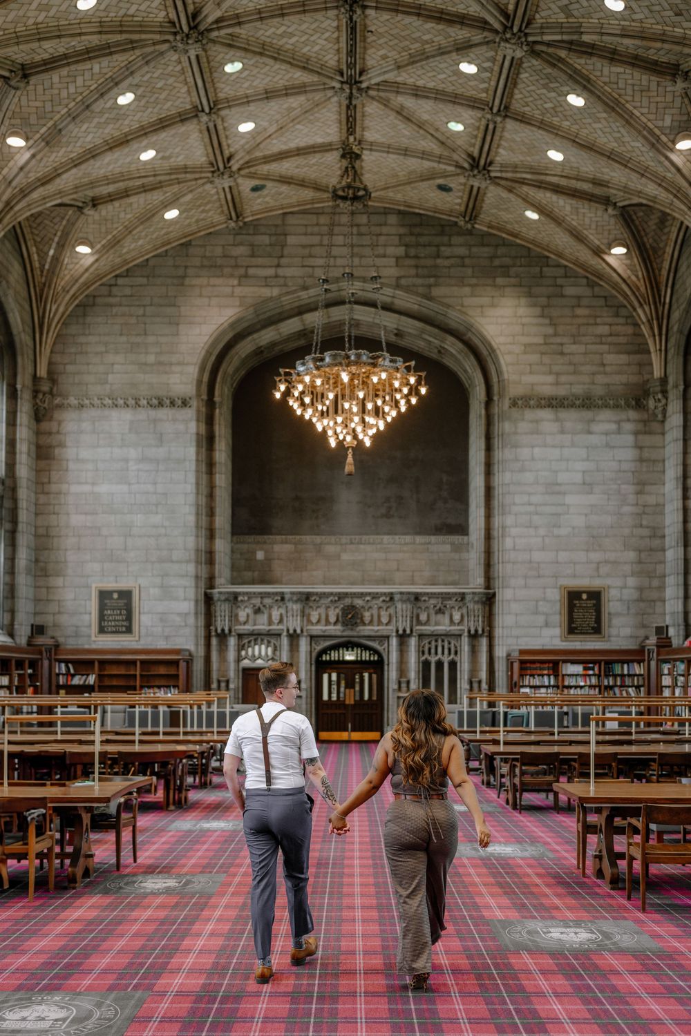 A couple walks hand in hand through a Gothic library hall with vaulted ceilings and a crystal chandelier.