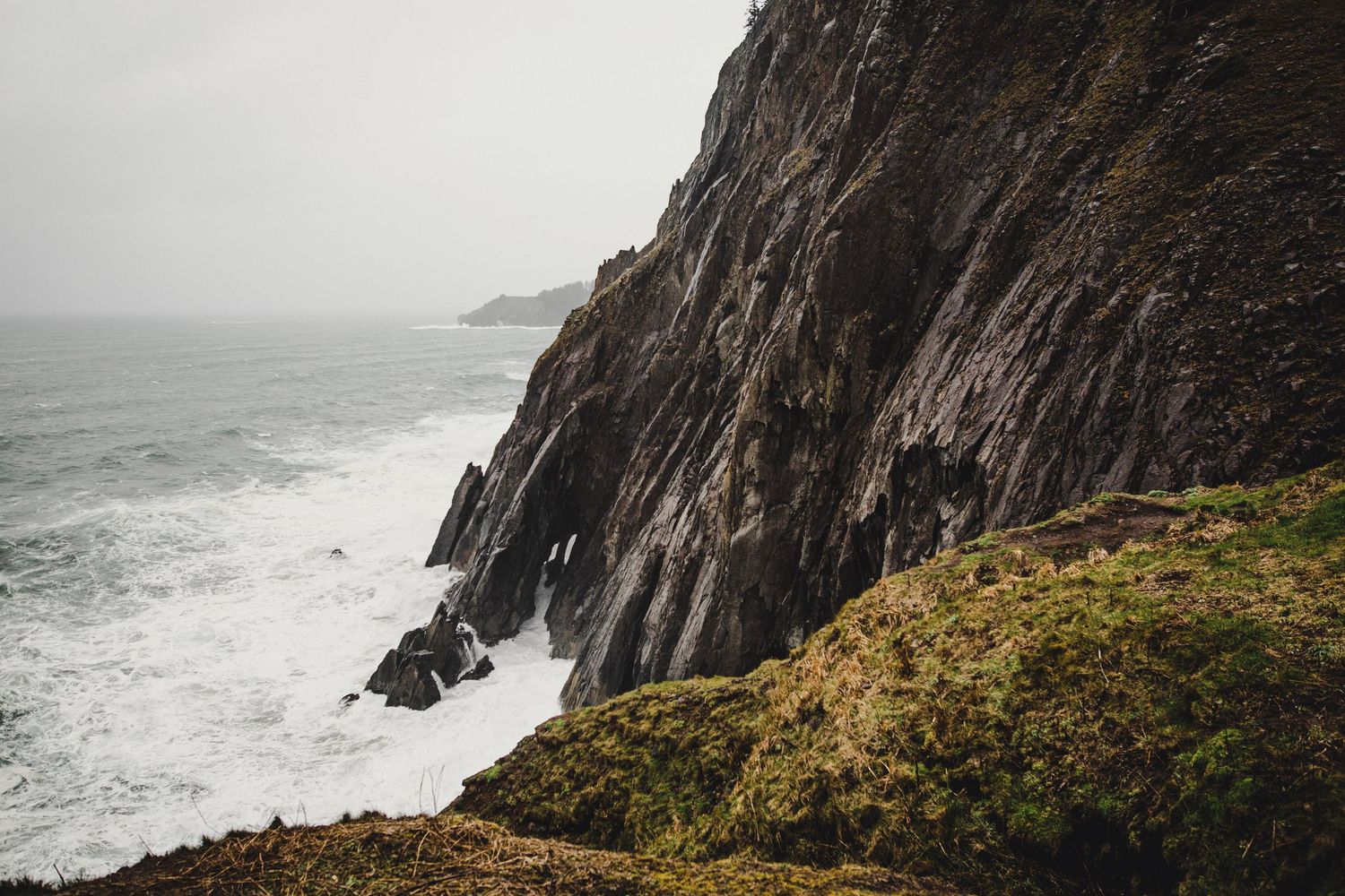 Dramatic rocky cliffs tower over crashing waves on a misty coastal shoreline with rugged terrain and ocean spray.