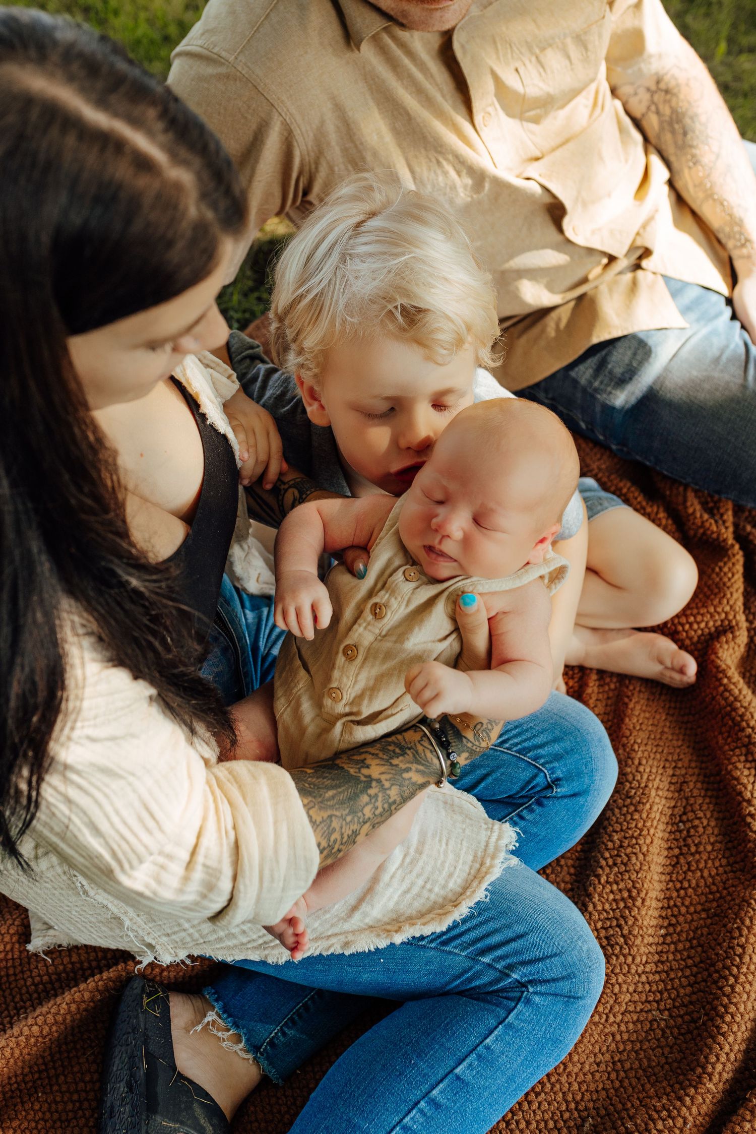 A tender family moment captured outdoors on a blanket as children share affectionate interactions while sitting together.