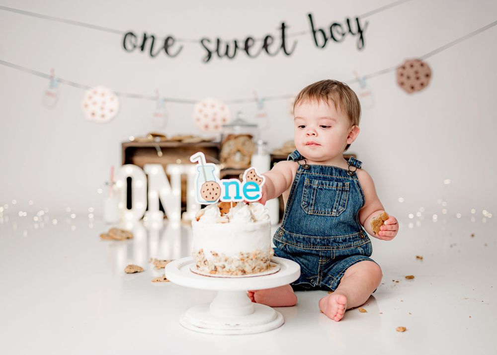 First birthday celebration scene with white cake, denim overalls, and decorative bunting and flowers on white backdrop.