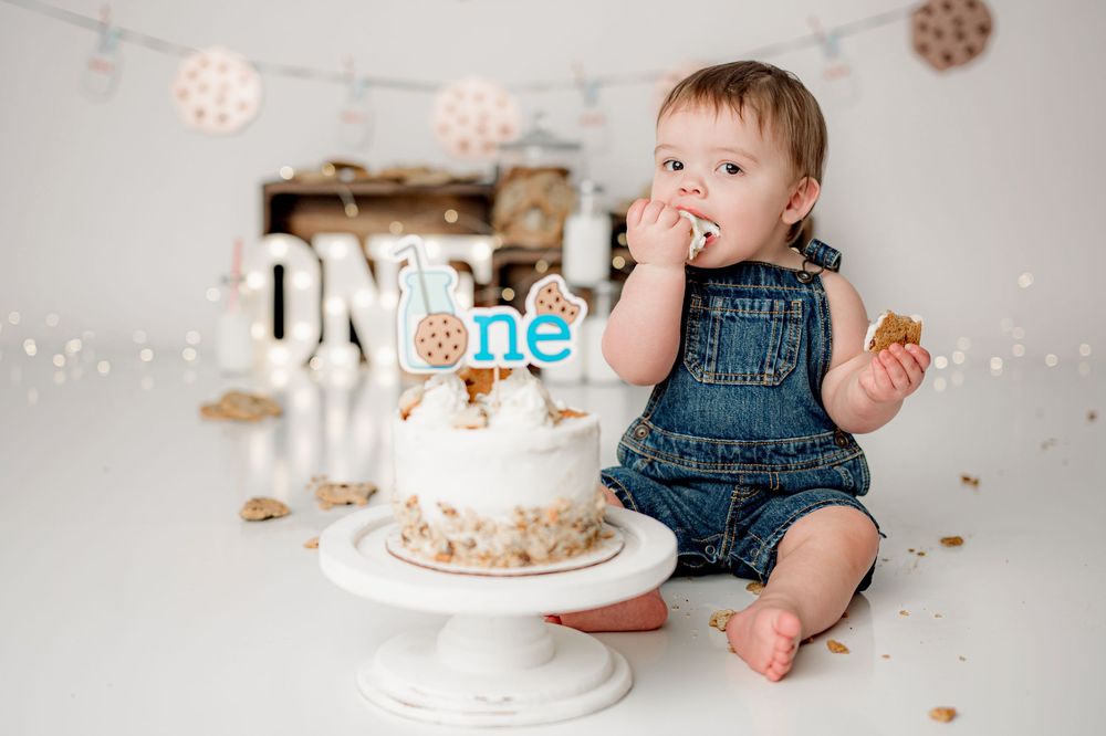 A baby in denim overalls sits on the floor beside a decorated first birthday cake with white frosting and string lights overhead.