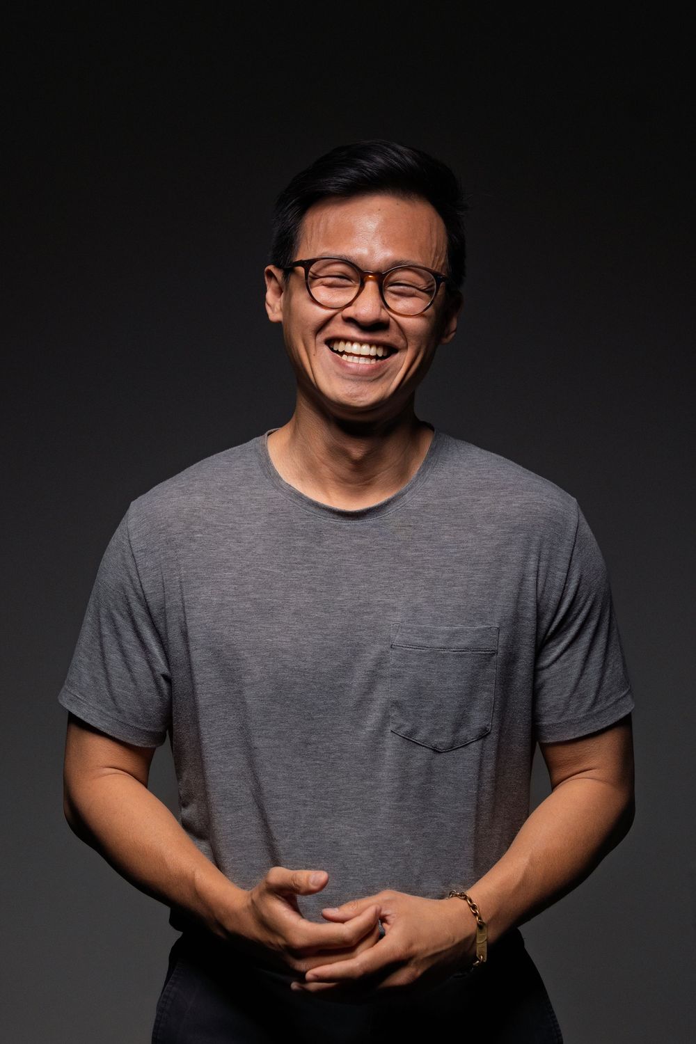 Portrait of someone wearing a gray t-shirt and glasses with a cheerful expression against a dark background.