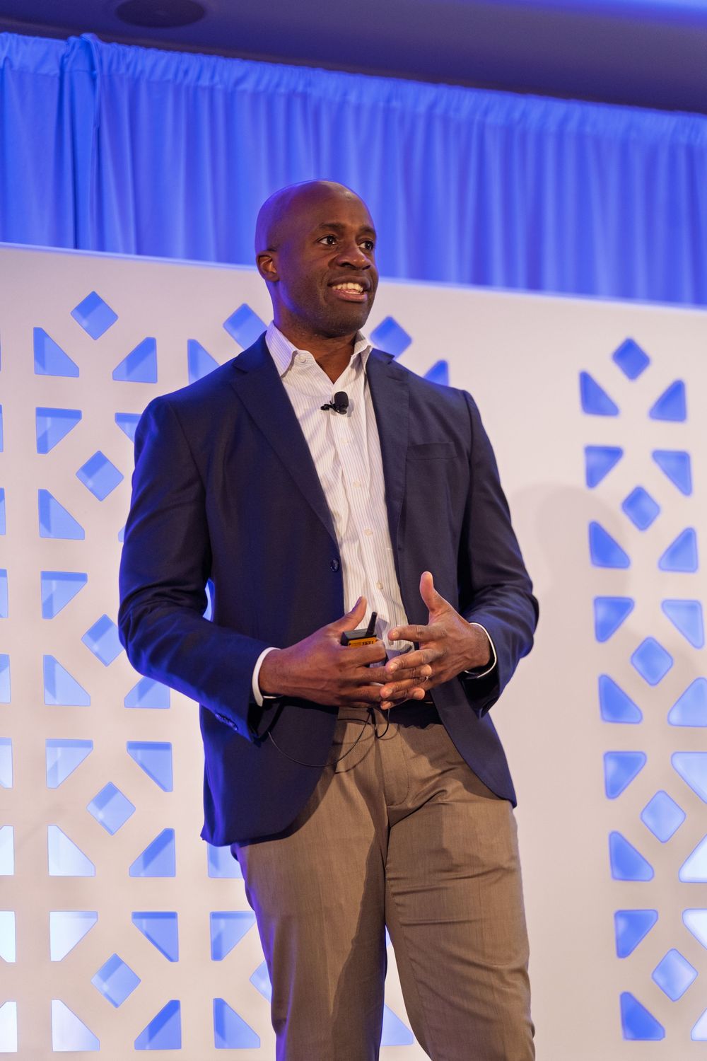 Professional person in navy blazer and khaki pants speaking on stage with white geometric backdrop and blue lighting.