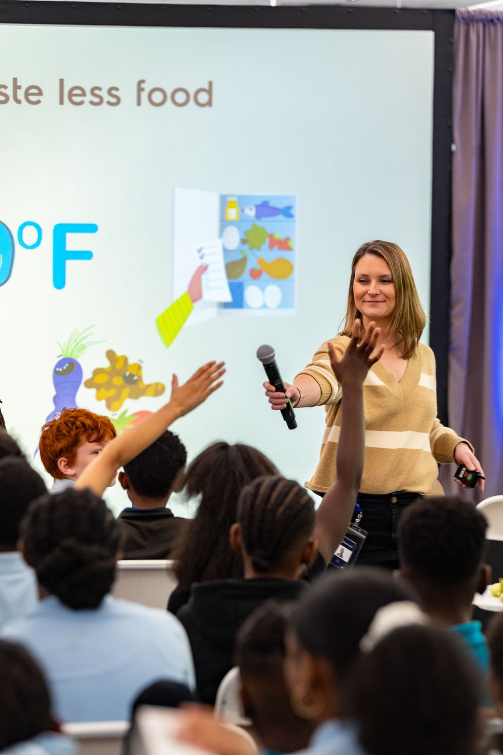 Students raise hands during interactive classroom lesson with presentation screen showing food waste information.