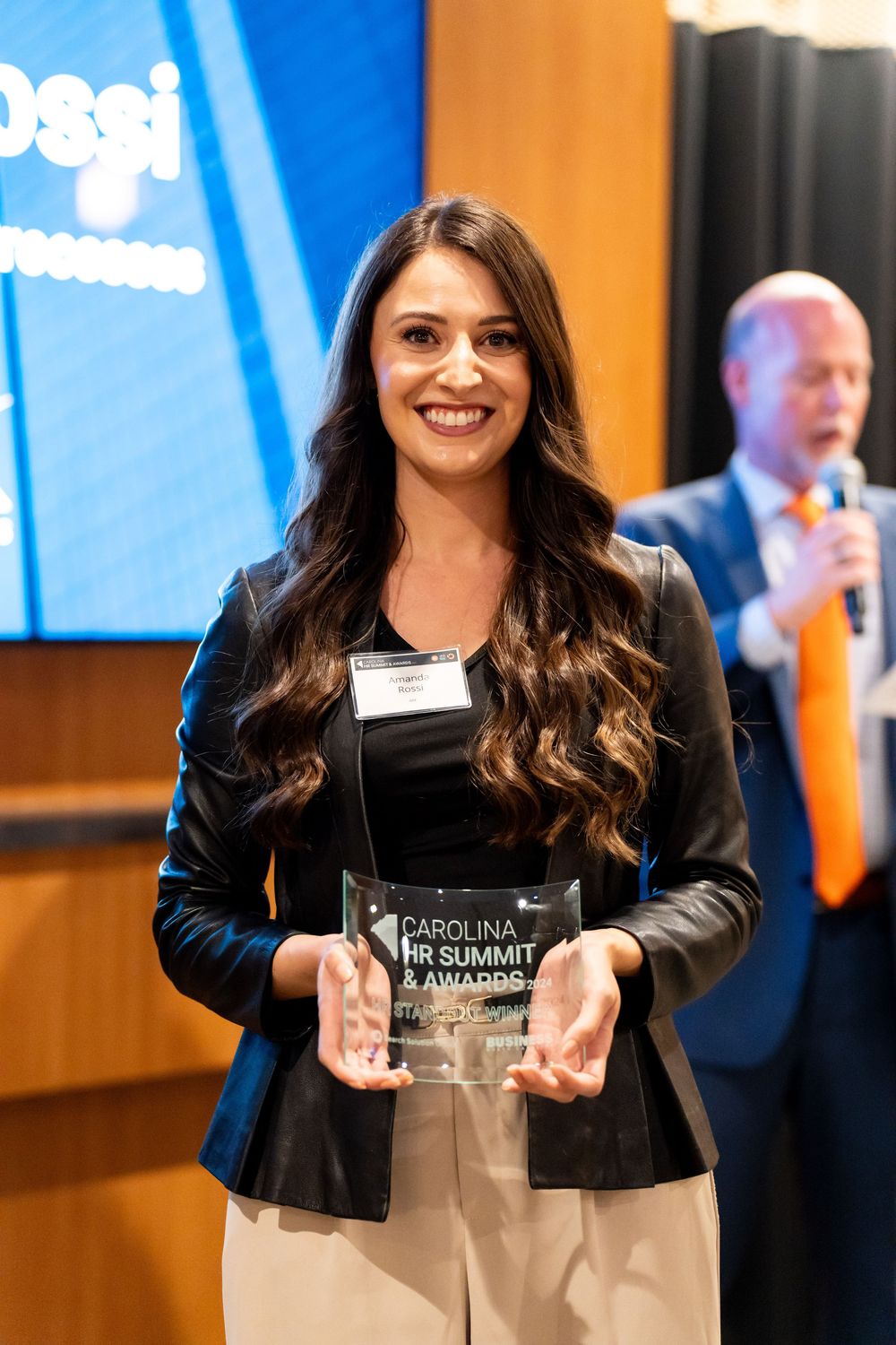 Professional event attendee in black attire receives an award and shows joyful expressions at an industry summit ceremony.