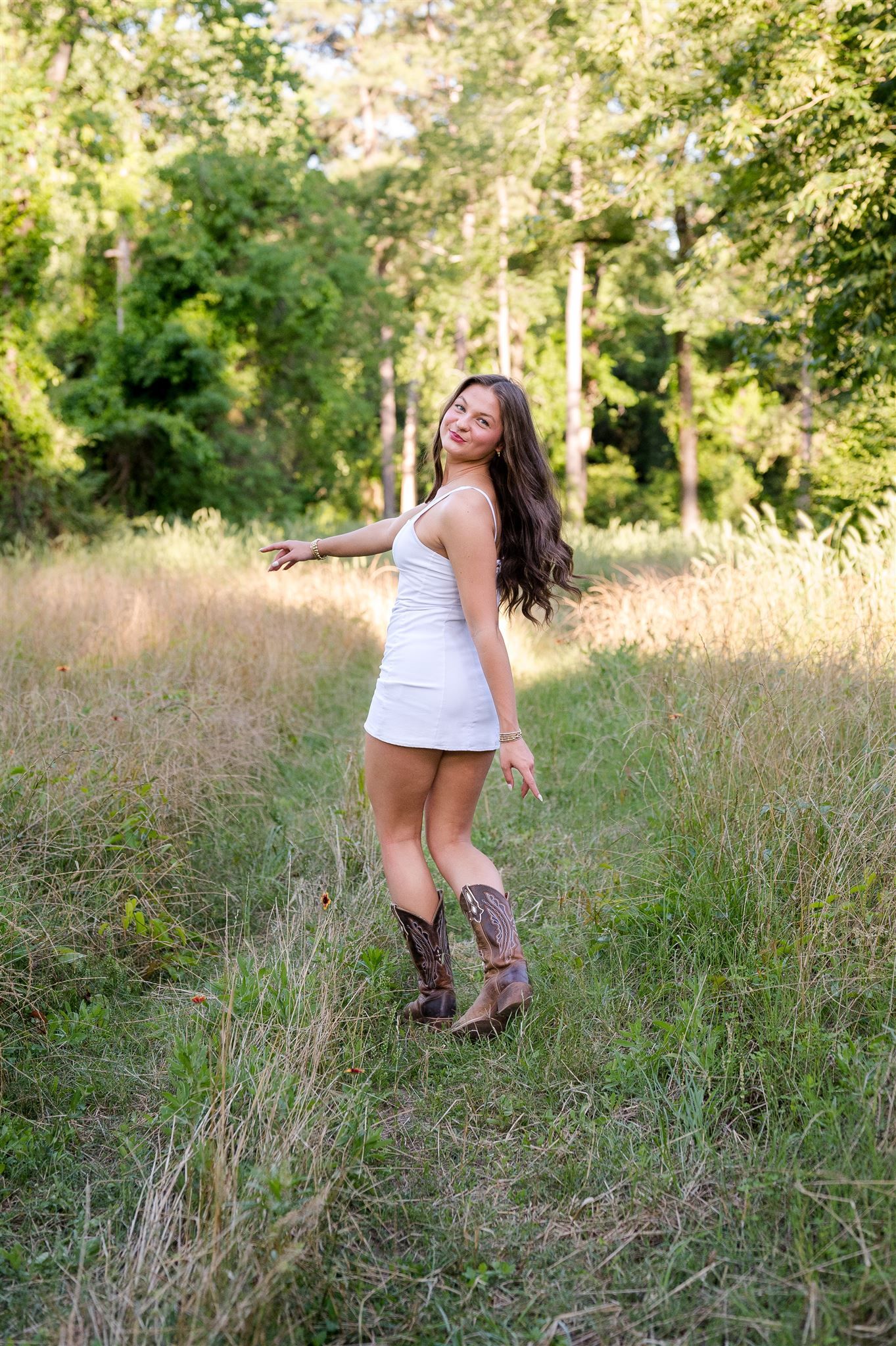 A white dress and cowboy boots stand out against a sunny meadow backdrop with tall grass and trees.