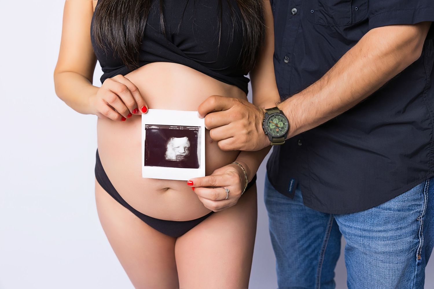A couple holds an ultrasound photo against a pregnant belly while wearing dark clothing against a white background.