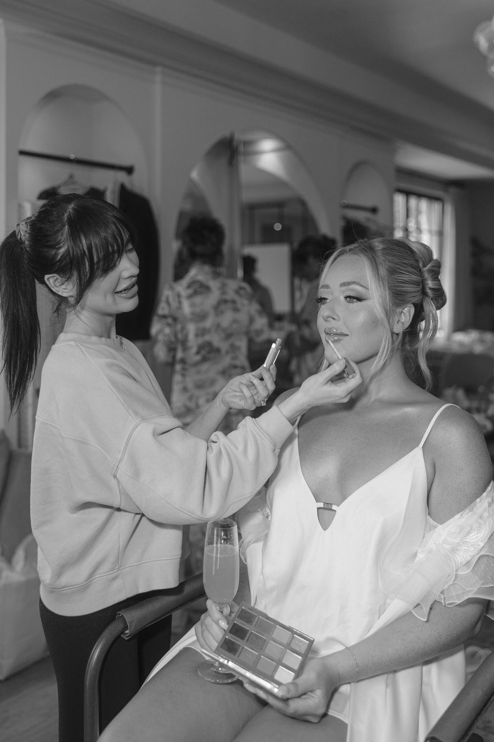 A makeup artist applies cosmetics to a bride in a white dress holding a makeup palette while getting ready.