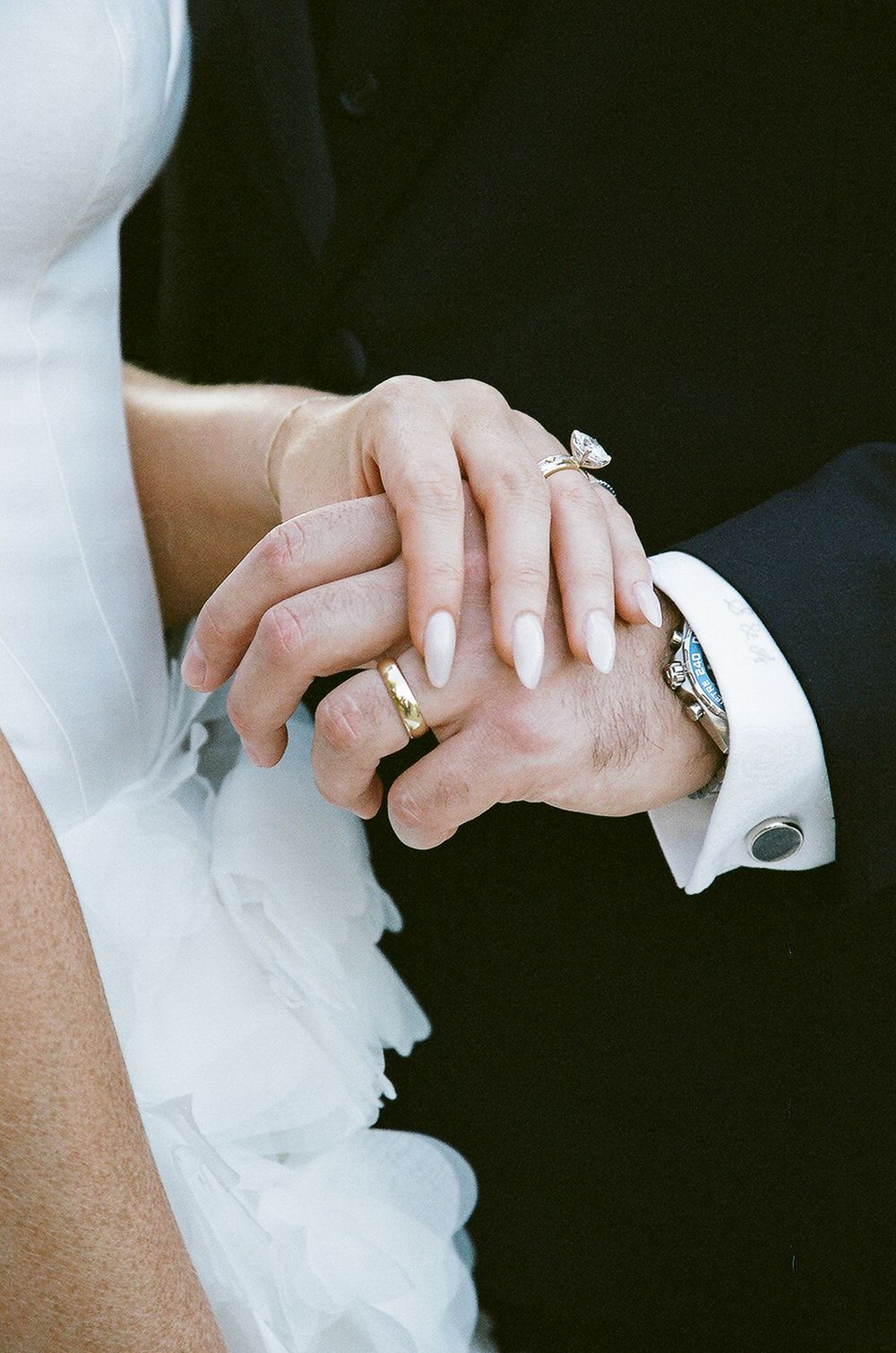 A close-up shot of newlywed hands showing wedding rings, with the bride in white and groom in dark formal attire.
