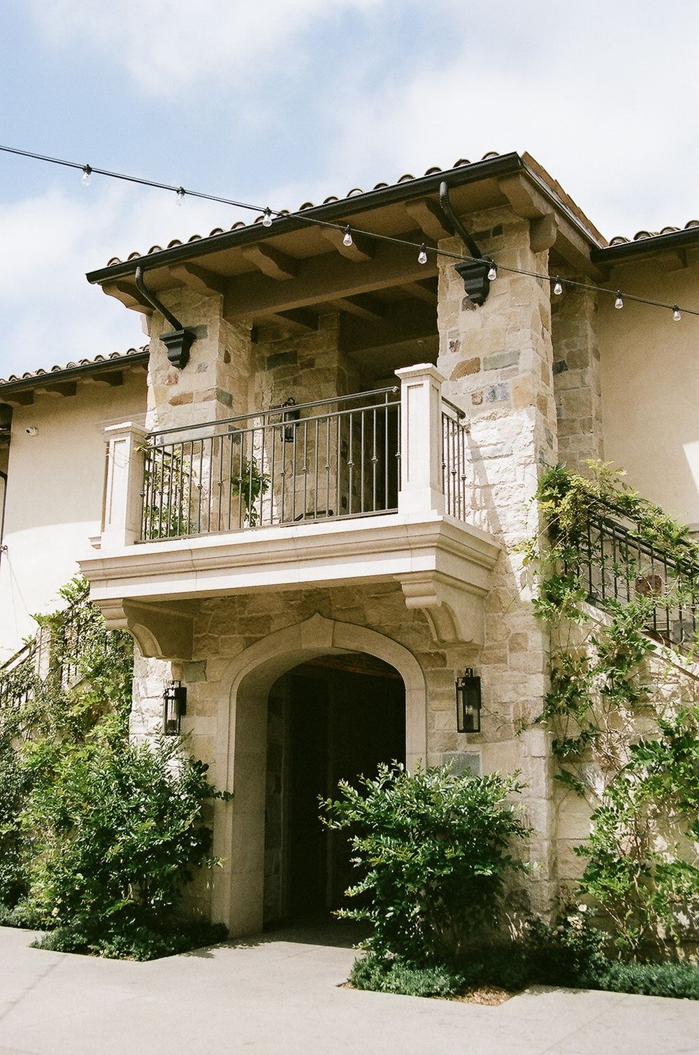 Mediterranean style stone archway entrance with wrought iron balcony and climbing vines on a white stucco exterior.