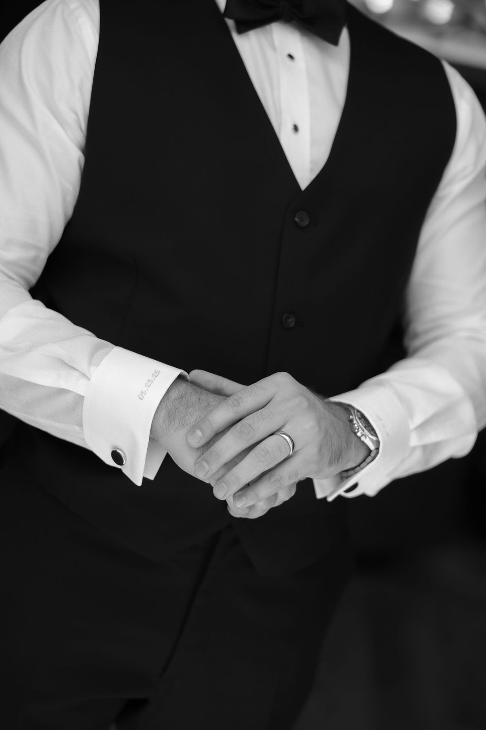 A black and white close-up shot of formal attire showing a black vest, white dress shirt, and hands adjusting cuff links.
