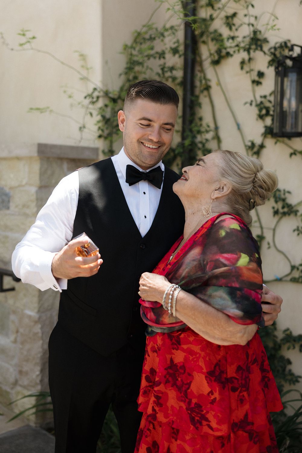 A couple shares a candid laughing moment together, with one wearing a tuxedo and the other in a red floral dress.