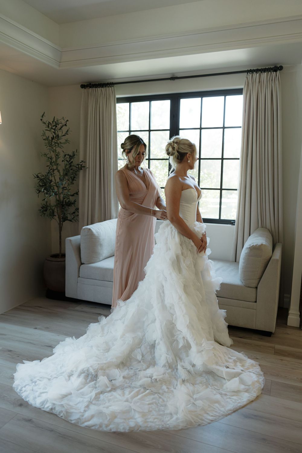 Two figures stand near a window as one wears an elegant white wedding gown with a flowing lace train.