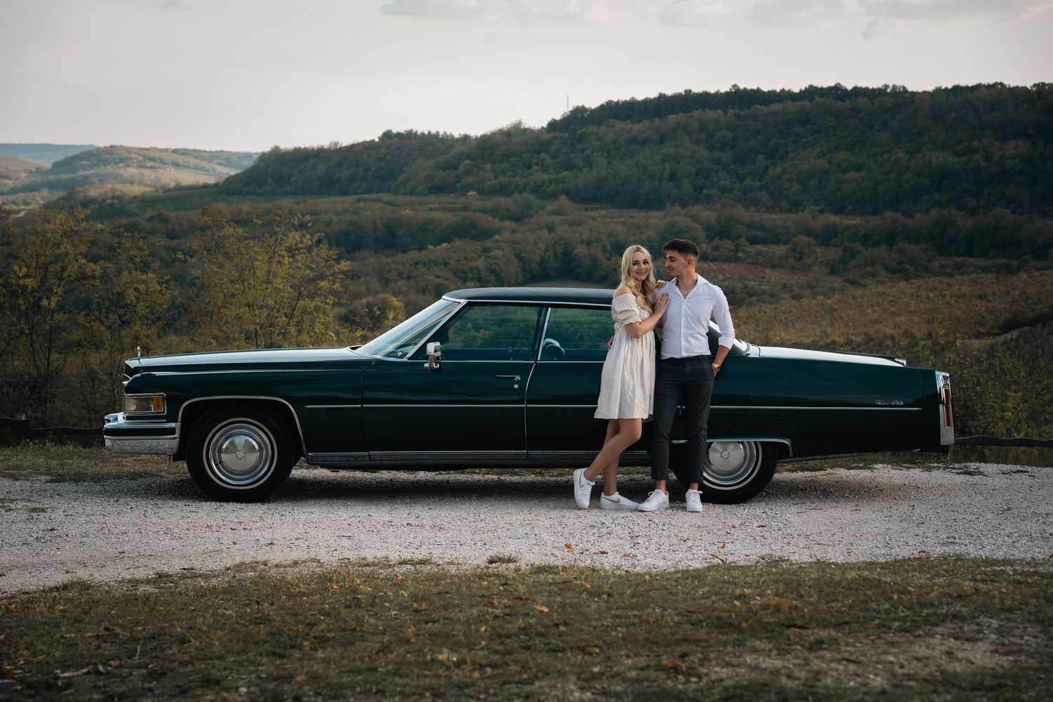 A classic dark Cadillac parked on a hilltop overlook with rolling mountains in the background.