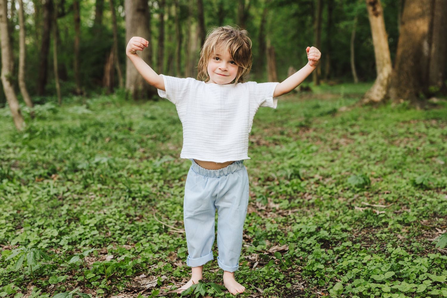 Child standing with raised arms in green forest clearing wearing white t-shirt and light blue pants.