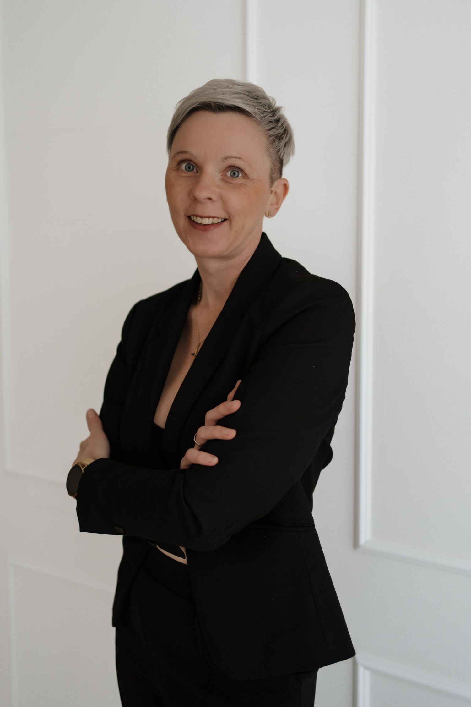 A professional stands confidently with arms crossed wearing a sleek black blazer against a white wall.