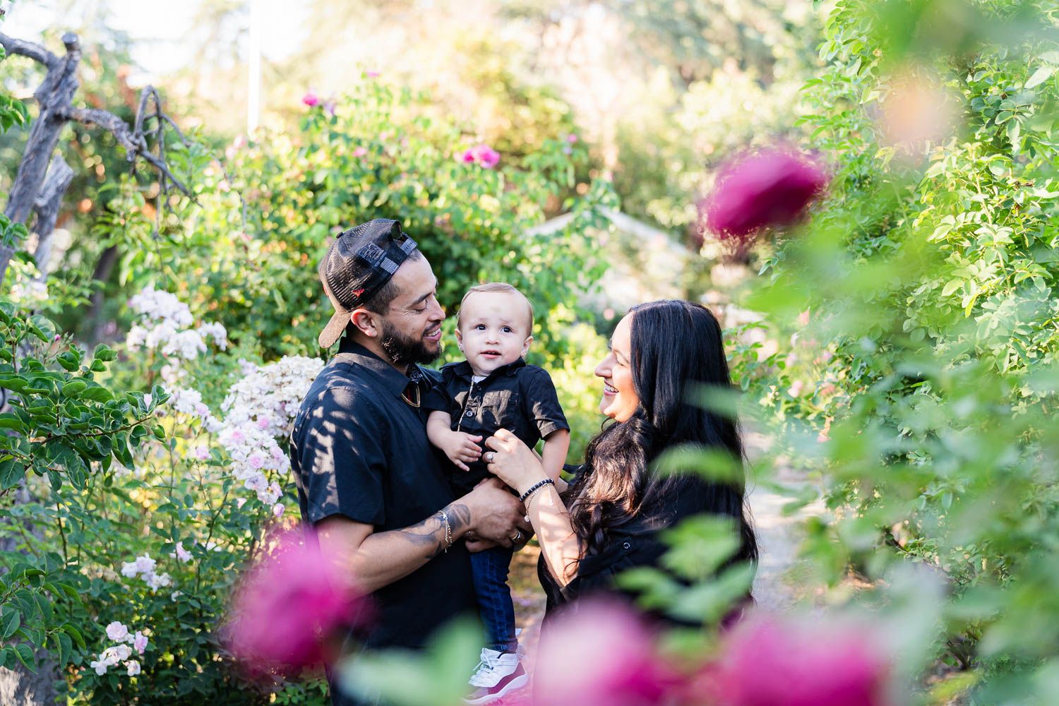 A family with young kid stand in a lush garden surrounded by blooming flowers and greenery on a sunny day.