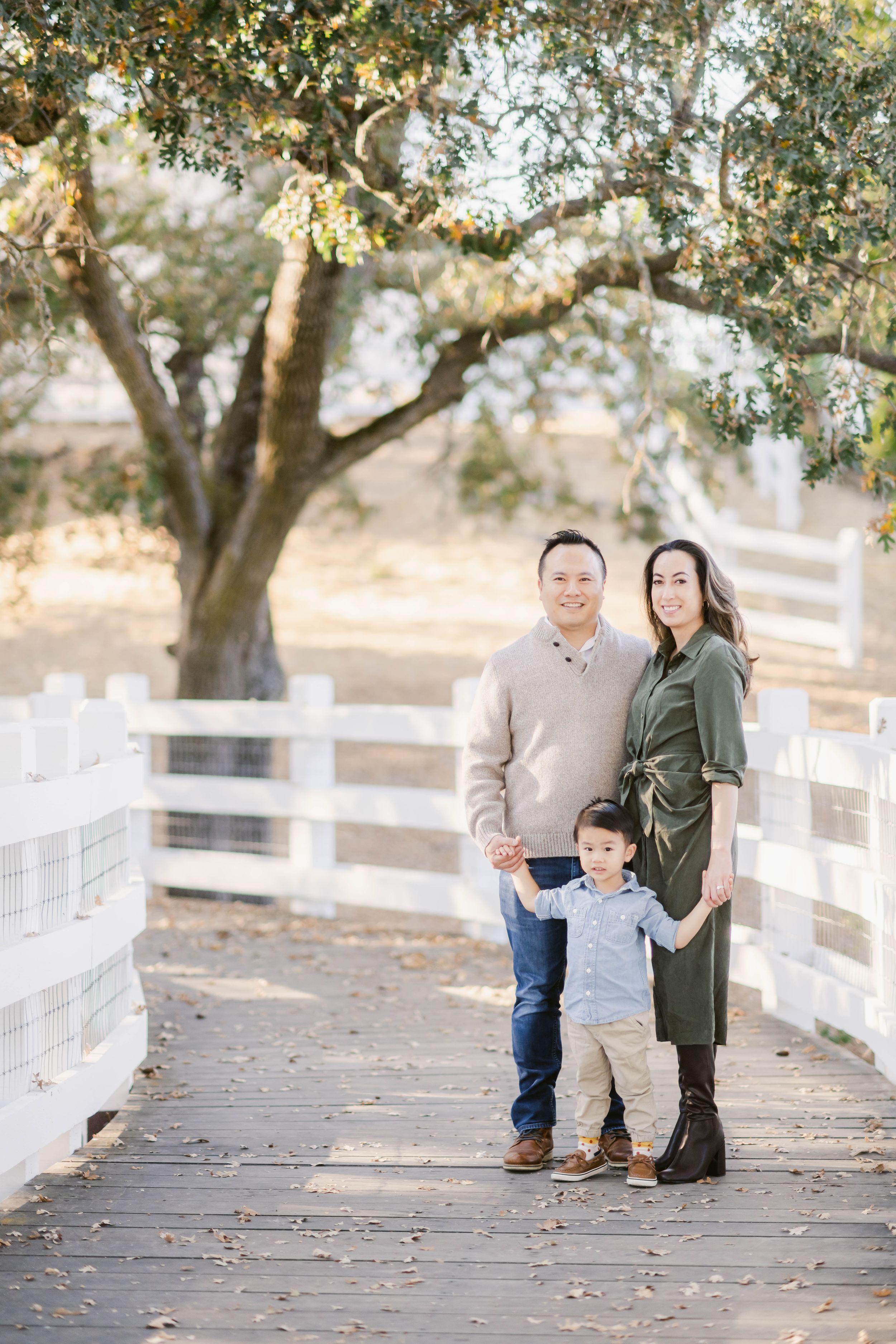 Family poses together outdoors on wooden boardwalk surrounded by oak trees and white fencing during autumn photo session.