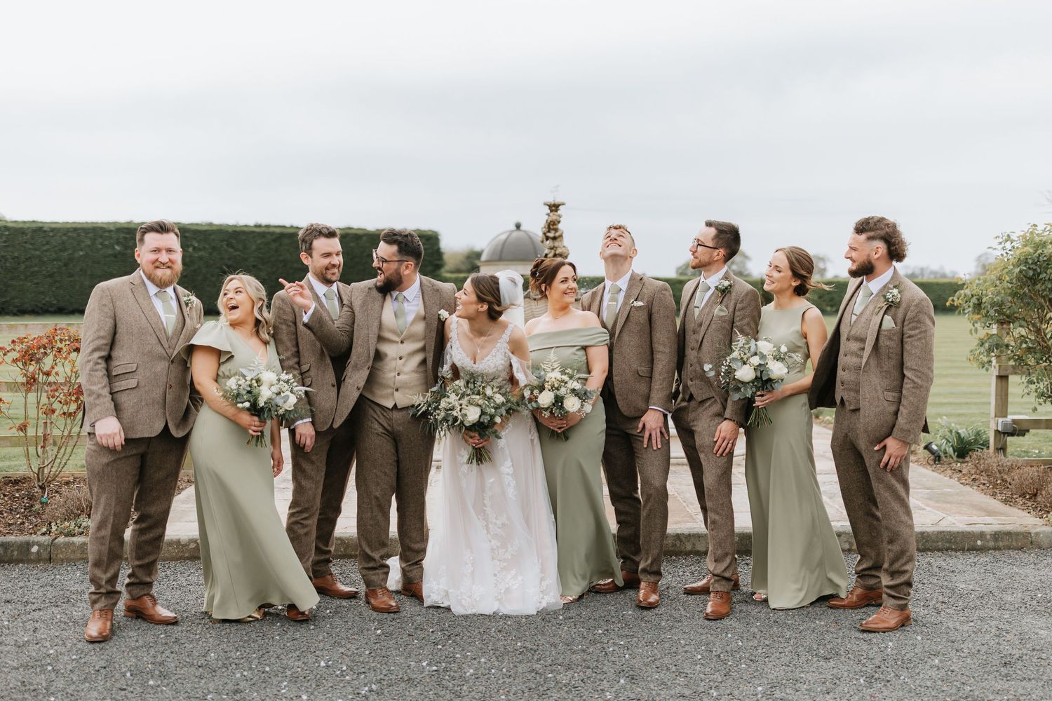Wedding party in sage green dresses and brown suits laughing together outdoors at a vineyard venue.