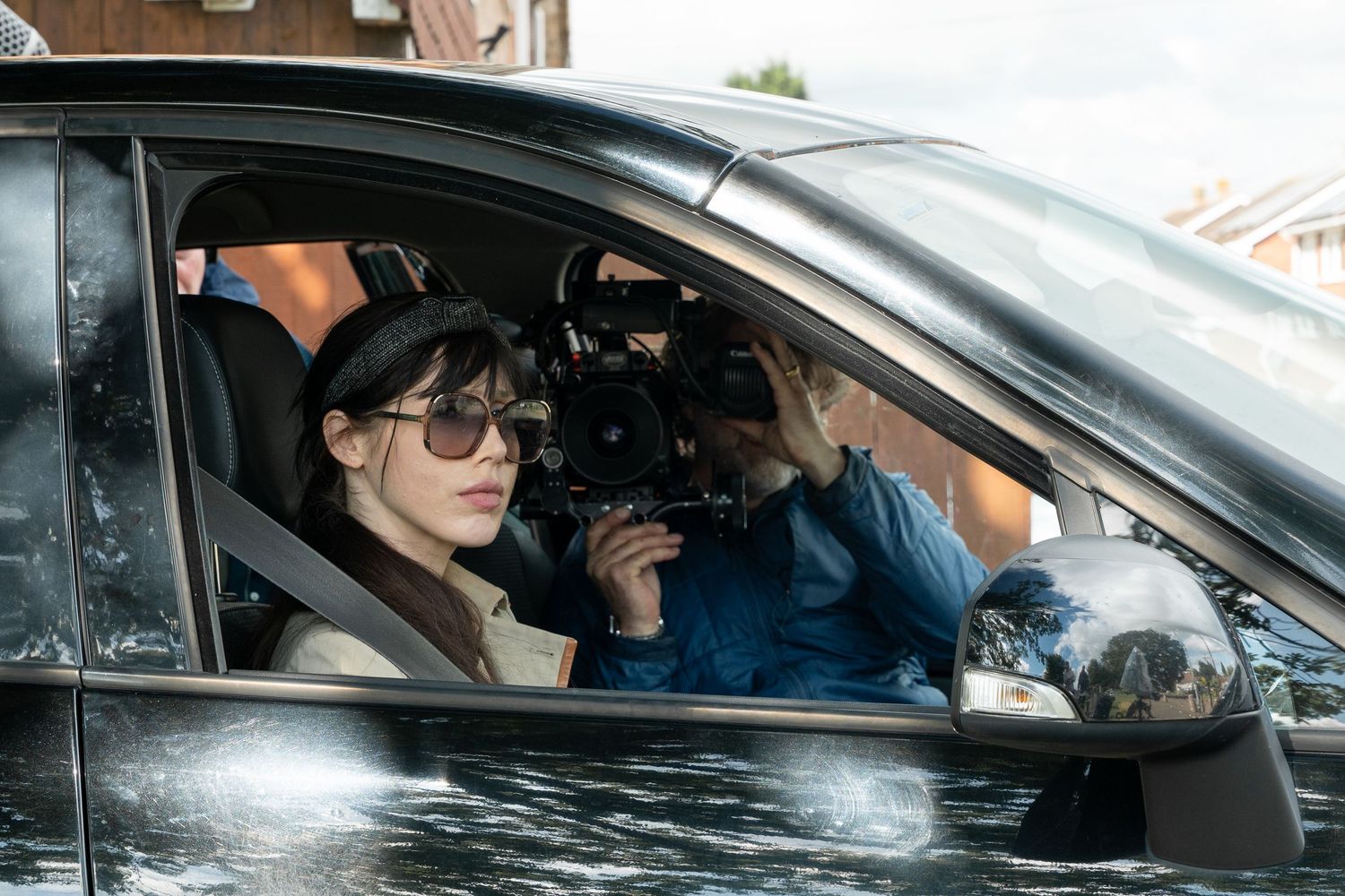 A TV crew films from inside a car as someone drives on a rainy day.