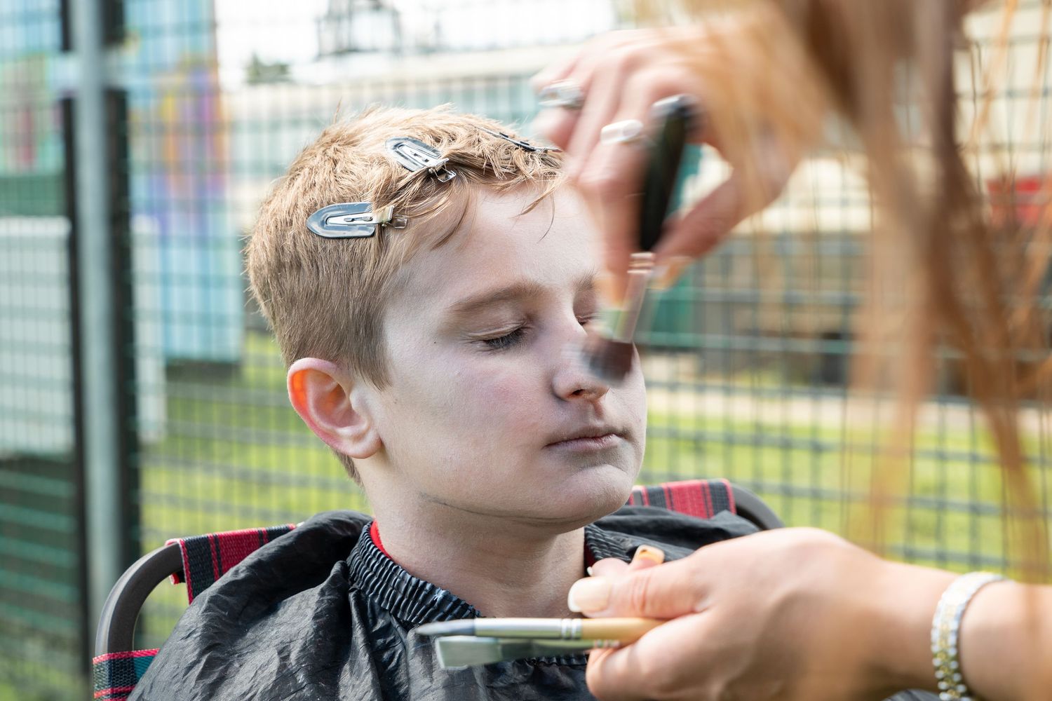 A child getting their hair trimmed outdoors with a haircut cape on while sitting in a chair at a playground area.