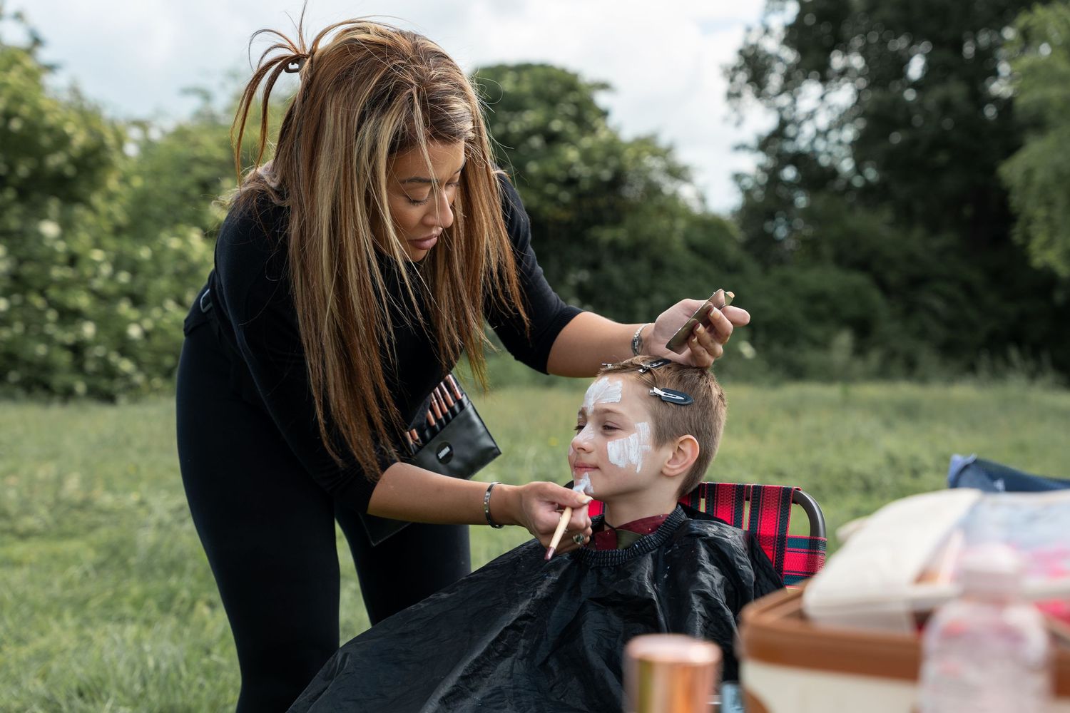 A makeup artist applies cosmetics to a young performer seated outdoors in a grassy area during daylight.