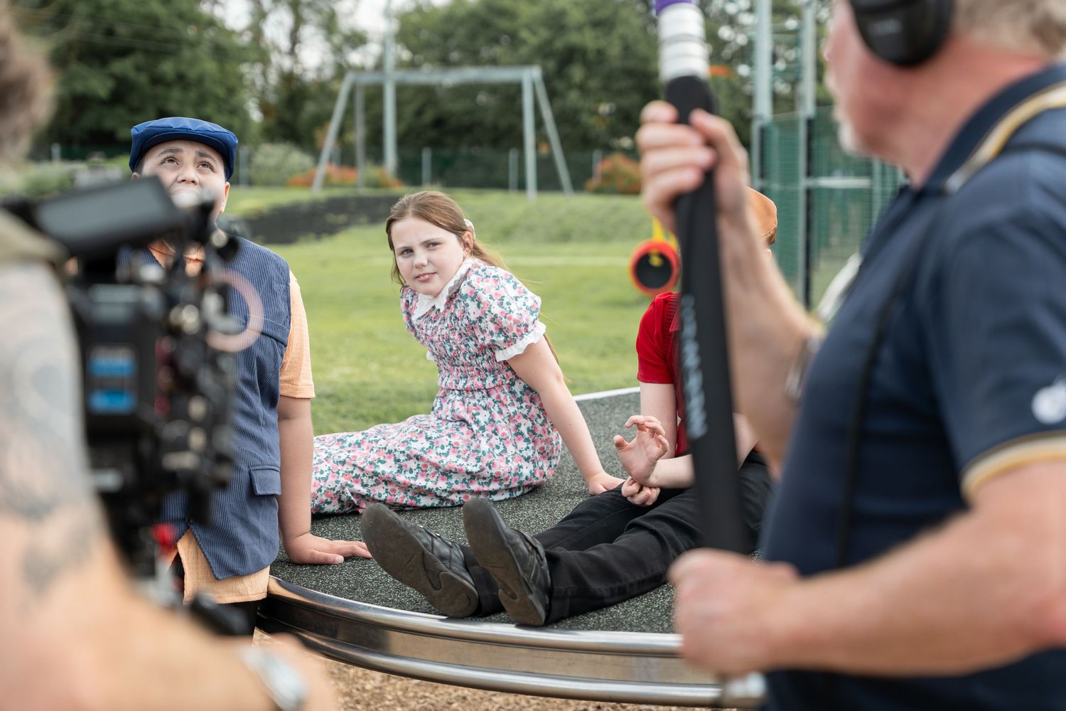 Film crew capturing scene of a person sitting on a dark table in a park setting while wearing a floral dress.