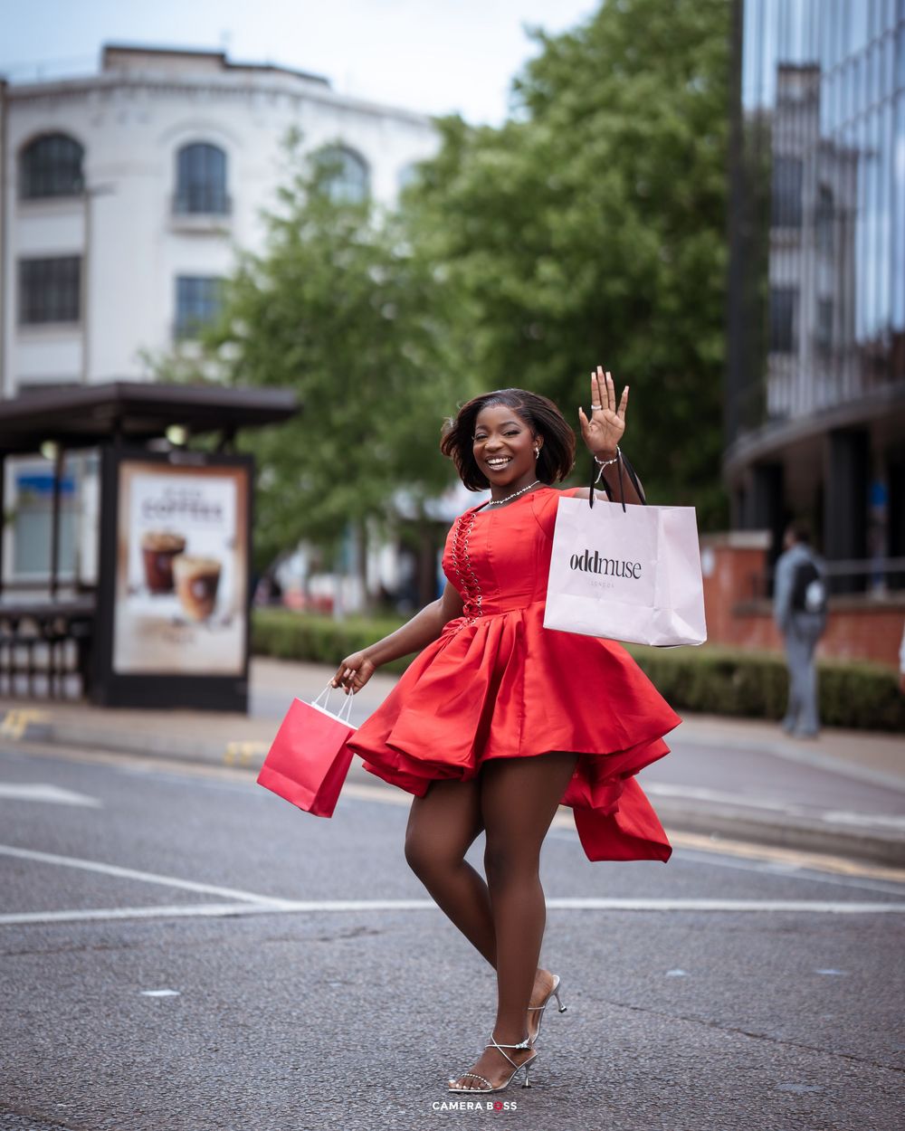 Joyful shopper in a flowing red dress twirls on a city street while holding shopping bags on a sunny day.