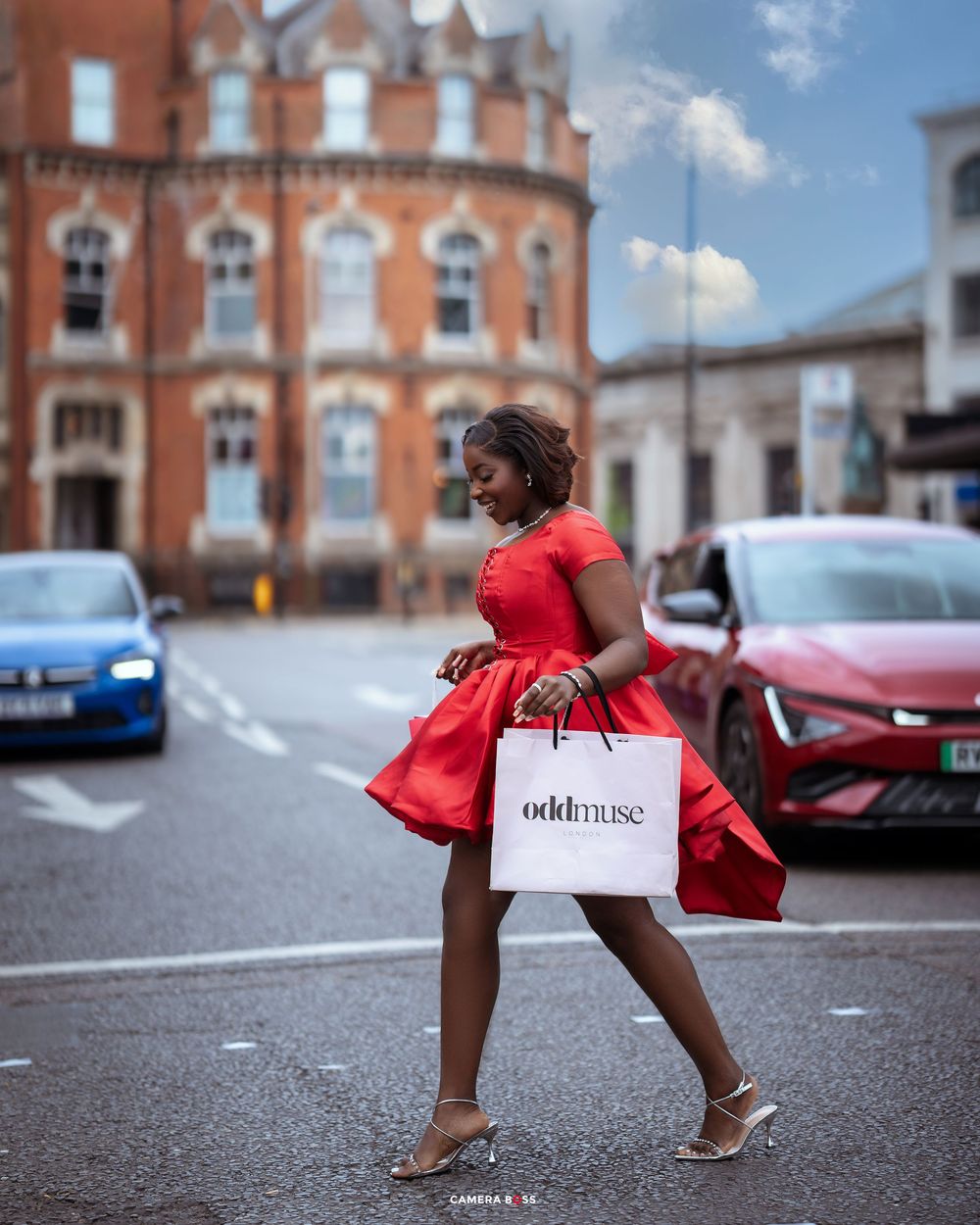 Person in a flowing red dress carries a shopping bag while crossing a street with historic architecture in the background.