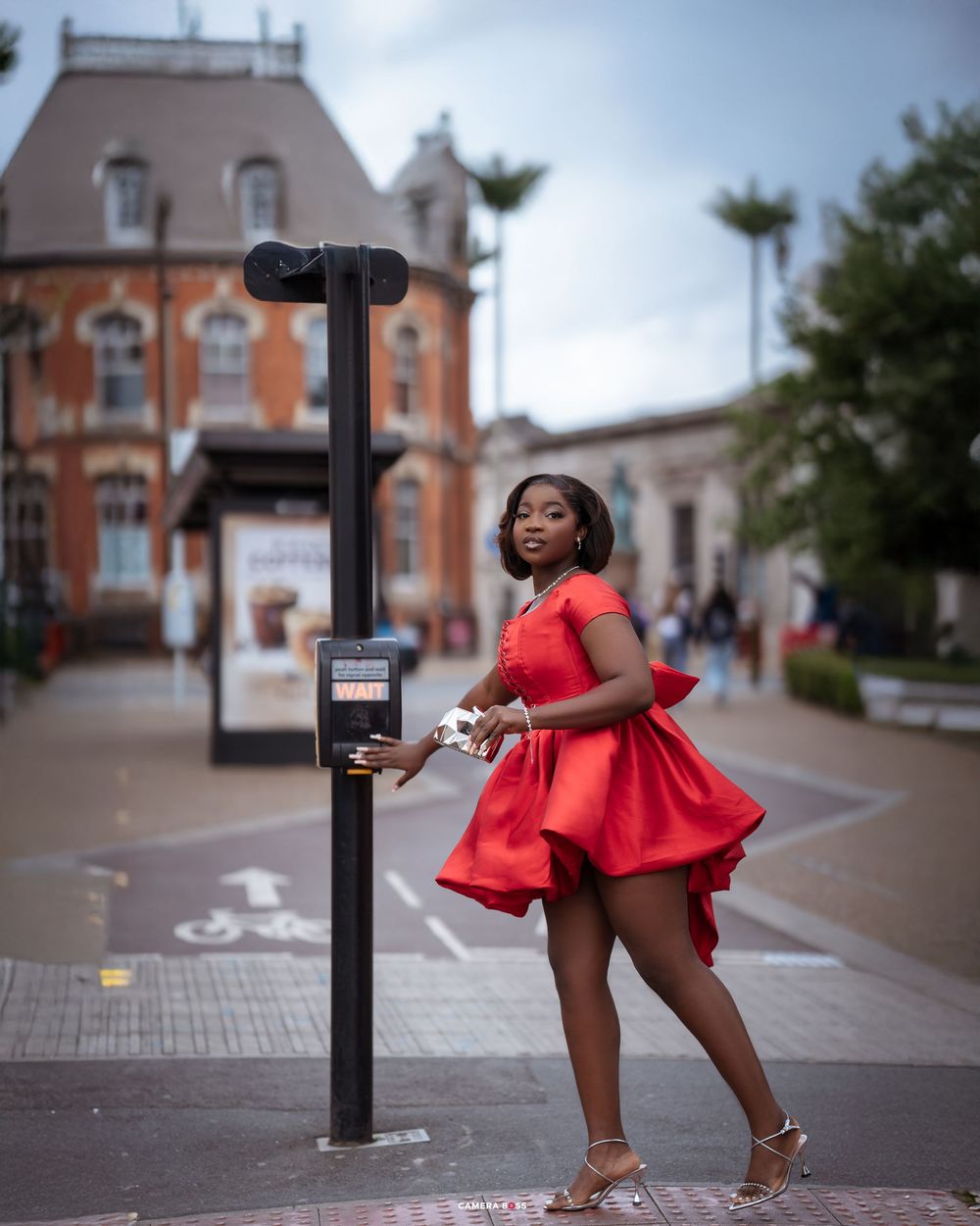 Person in a red dress poses playfully near a parking meter on a city sidewalk with brick buildings behind.