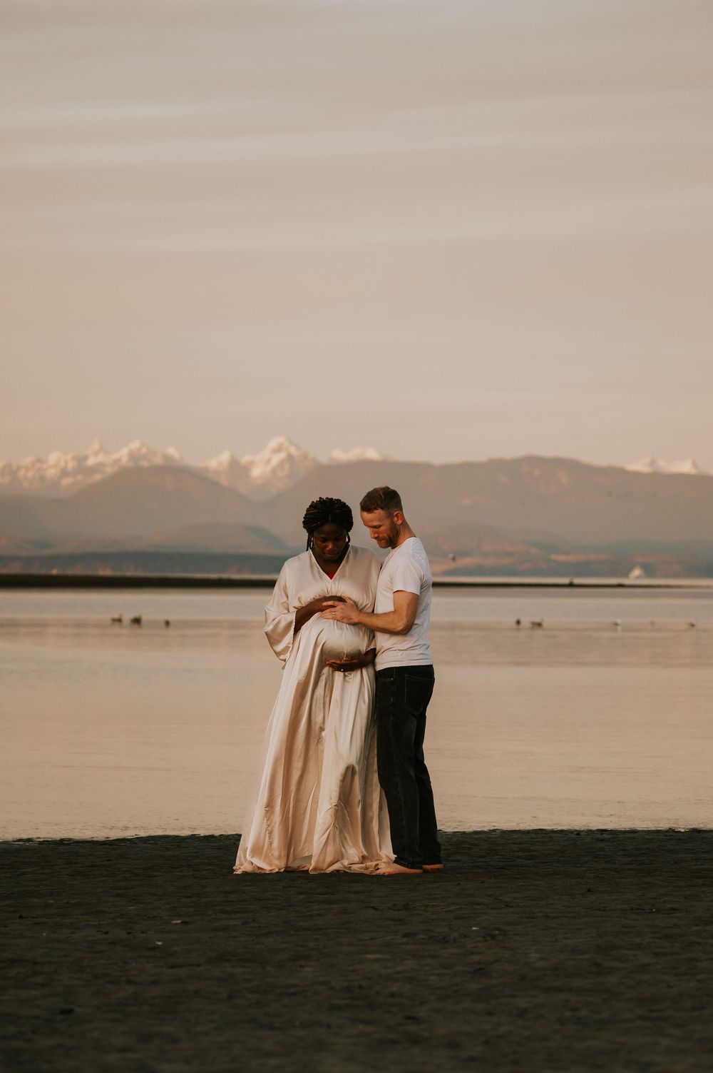 Maternity photography of couple holding bump at kye bay beach by ocean in front of mountains Comox Valley Vancouver Island photographer
