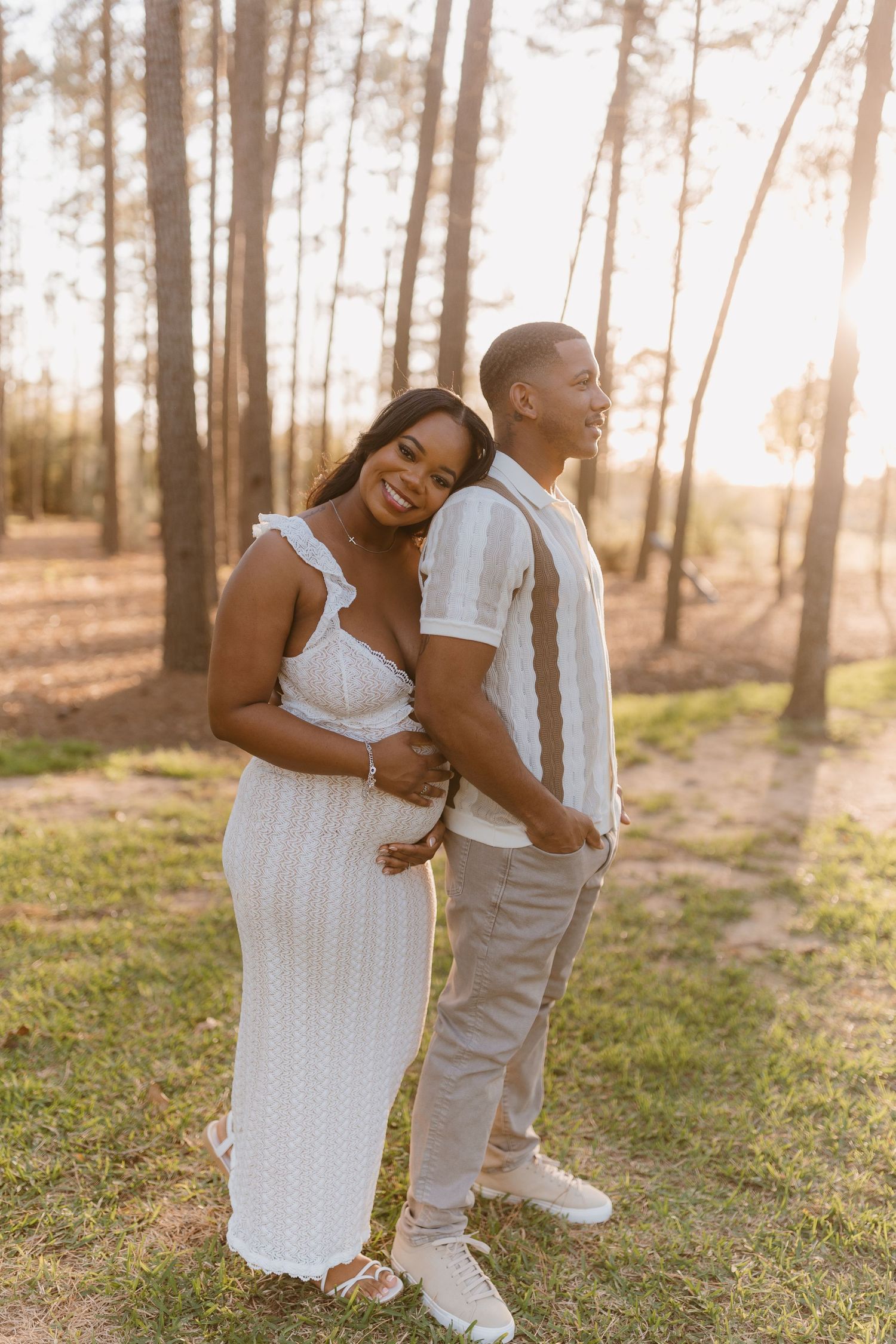 A couple in white outfits share a joyful moment together among tall pine trees at sunset.