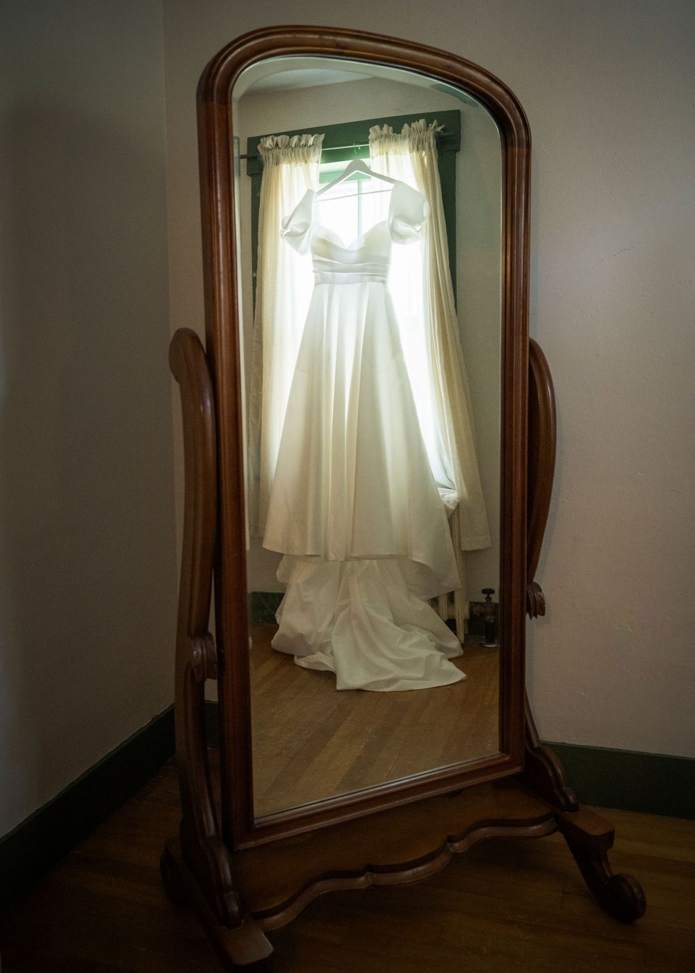 An elegant white wedding dress hangs in front of a vintage full-length wooden mirror in soft natural light.