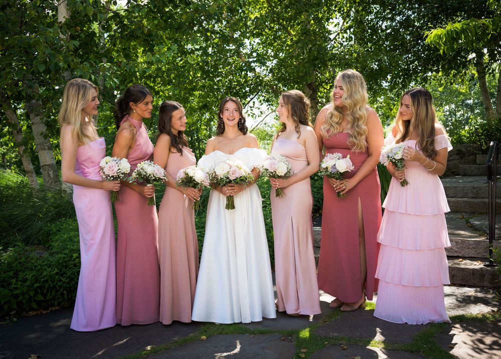 Bridal party in pink and mauve dresses poses outdoors in a garden setting with lush greenery.
