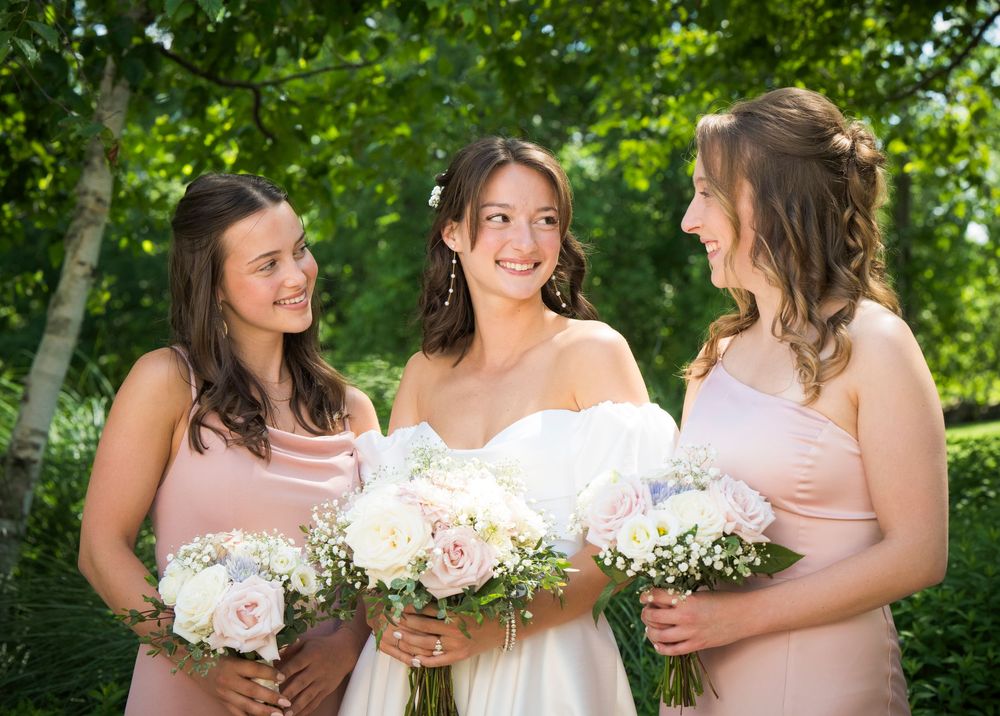 Three women in pink and white dresses hold beautiful floral bouquets while celebrating outdoors in a green garden setting.
