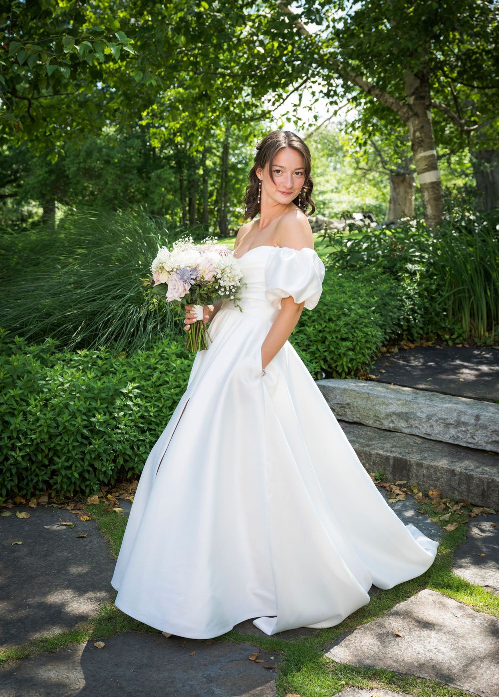 A person in a flowing white off-shoulder wedding gown poses with a bouquet in a lush garden setting on a summer day.